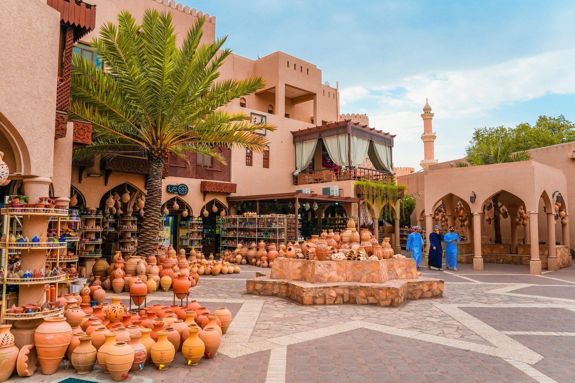 Vibrant market square with traditional buildings, a palm tree, numerous terracotta pots, and a distant minaret.