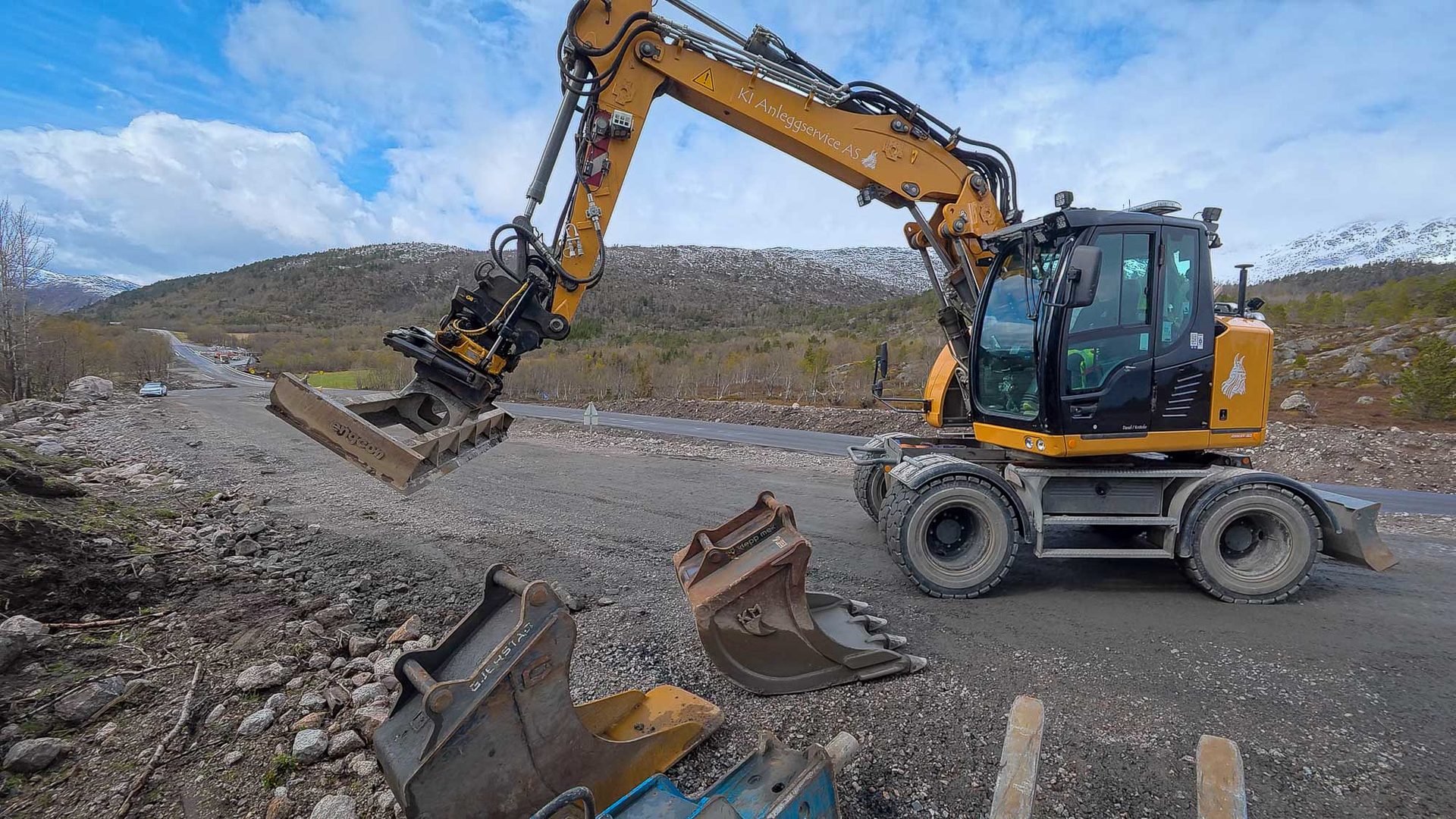Yellow wheeled excavator with grapple and buckets at a construction site by a road, mountains in background.