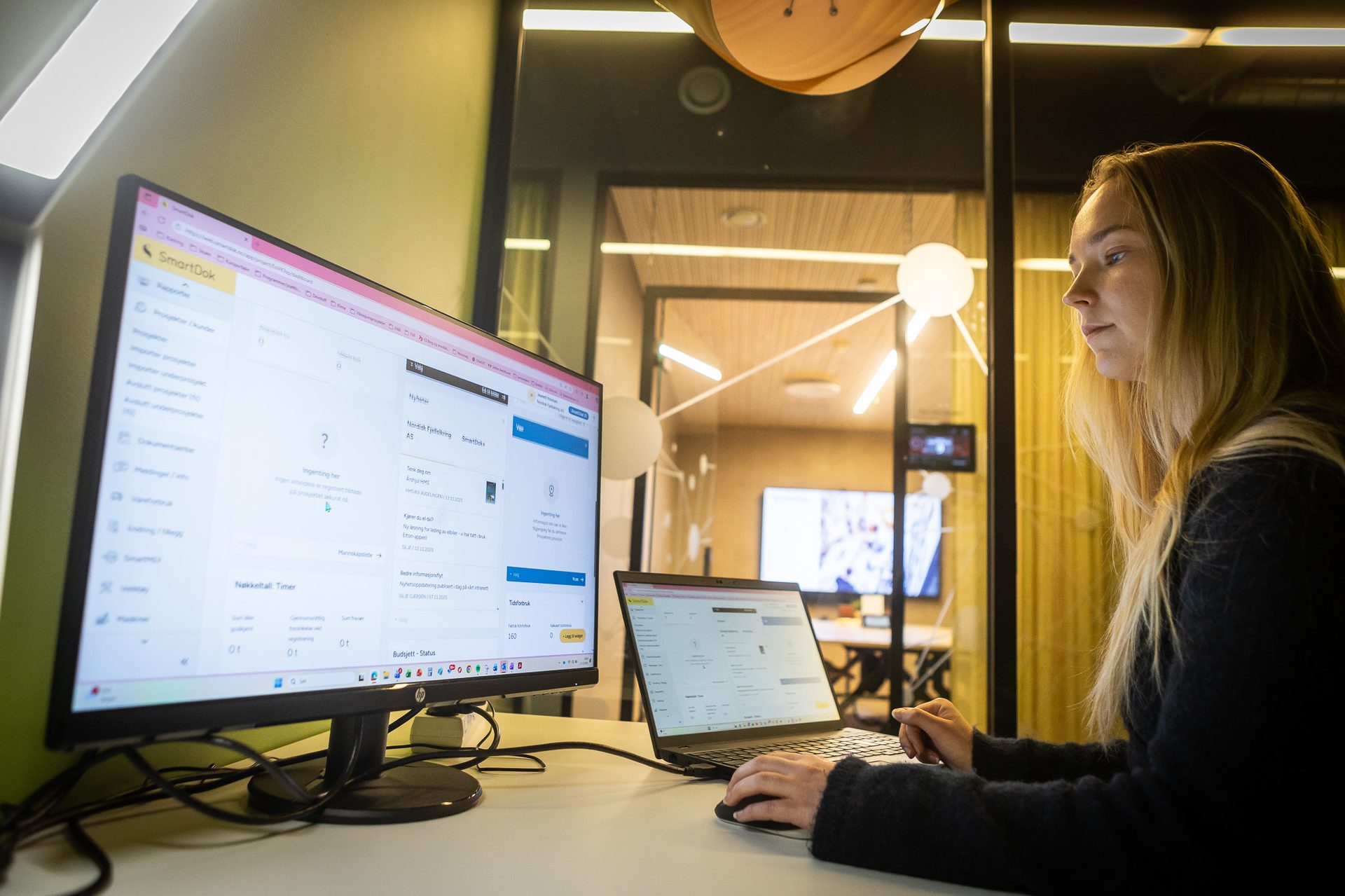 Young woman working at a desk with two computer screens showing "SmartDok" software in an office.