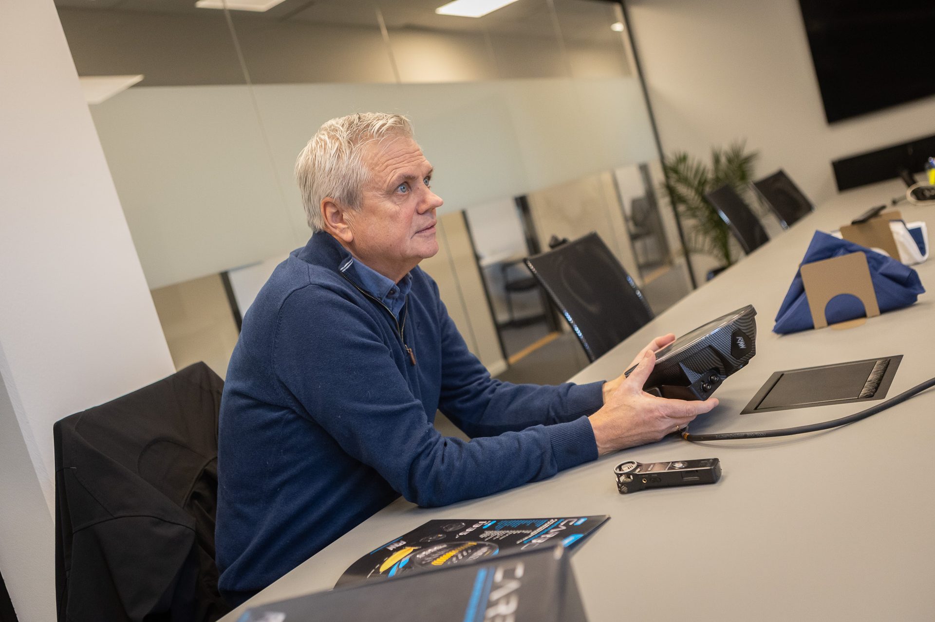 Man in blue sweater holds a black device labeled 'RAW' at a conference table, looking left.