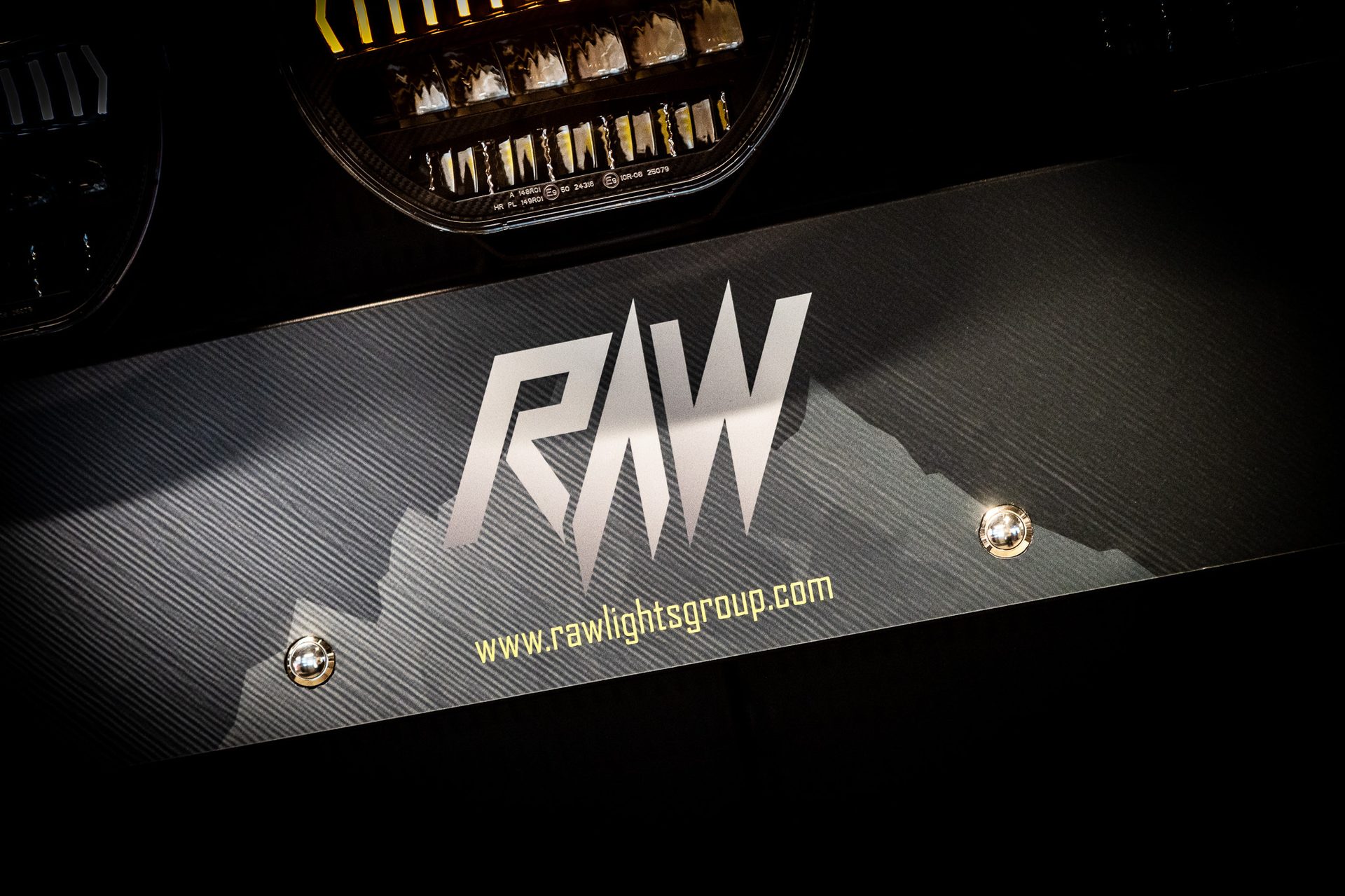Close-up: RAW logo & www.rawlightsgroup.com on dark textured panel, illuminated headlights above.