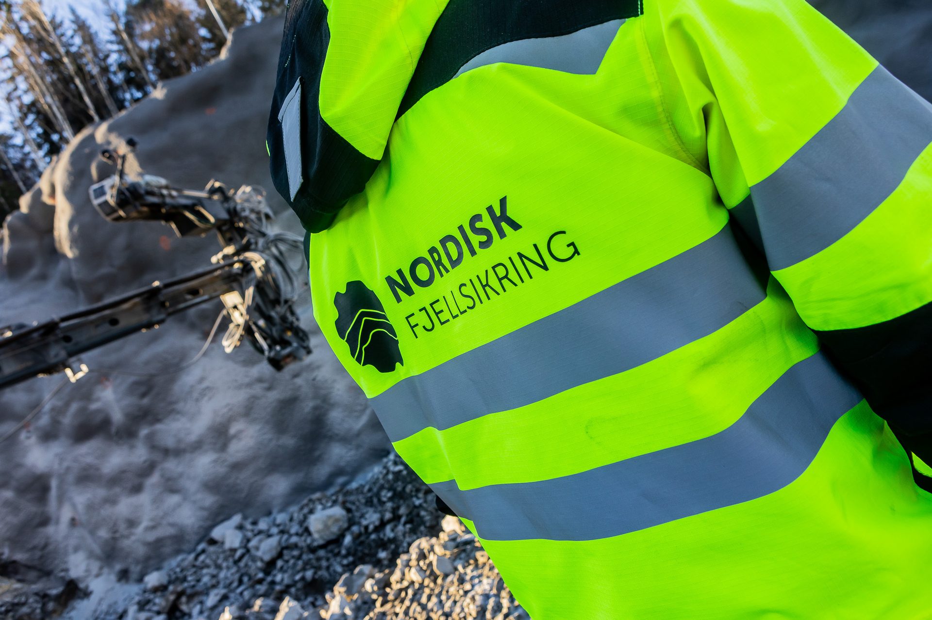 Back of person in a high-vis jacket with "NORDISK FJELLSIKRING" logo, near a drilling machine on rocky terrain.