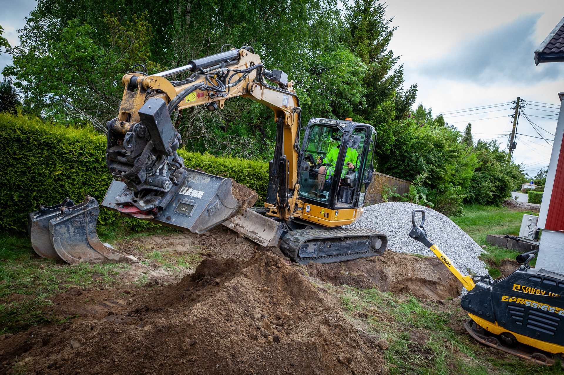 A person operates a mini-excavator, digging dirt next to gravel and a compactor.