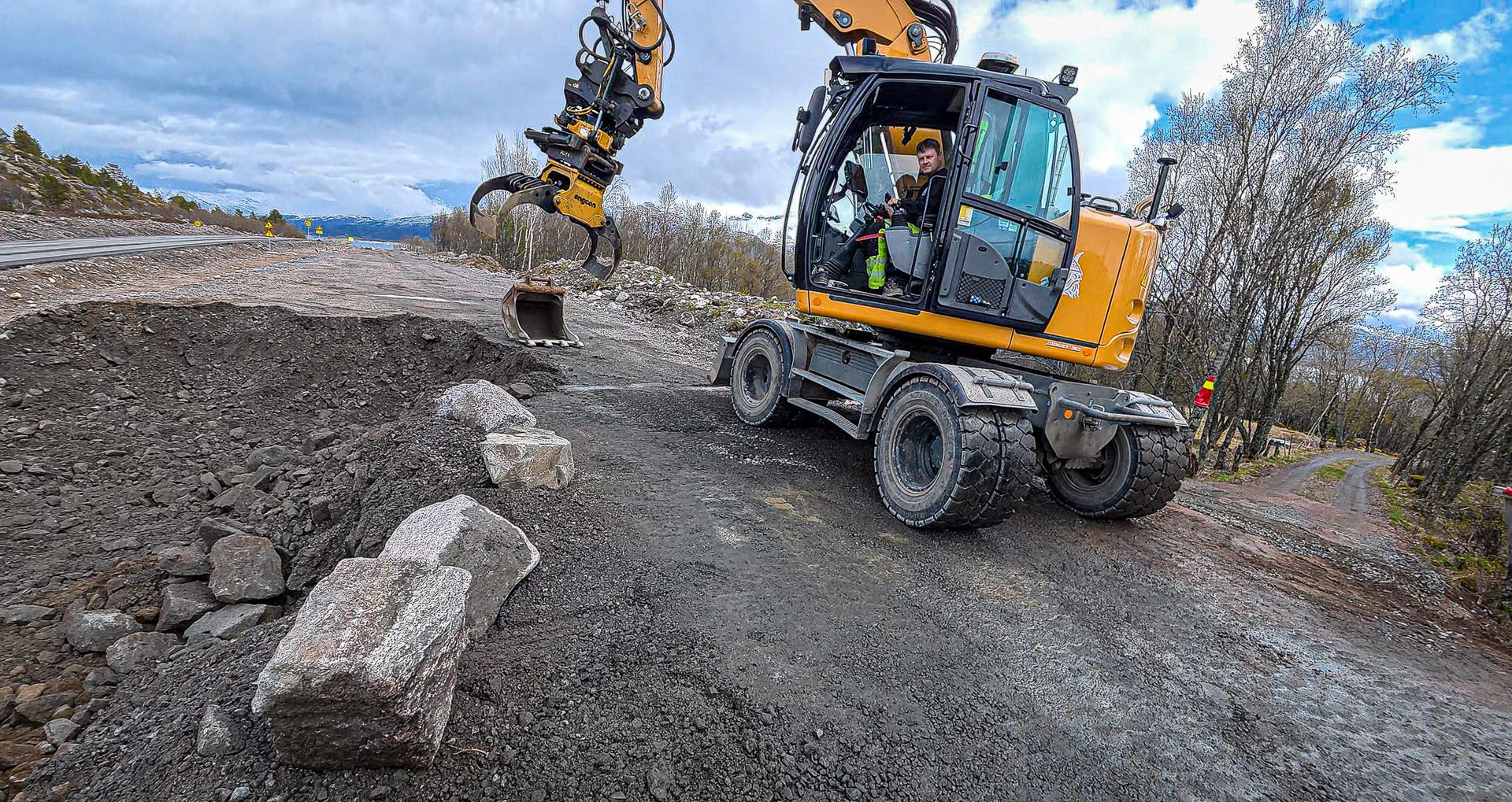 A yellow excavator with a grabber arm is working on a dirt road next to a large pile of earth, with mountains in the background.