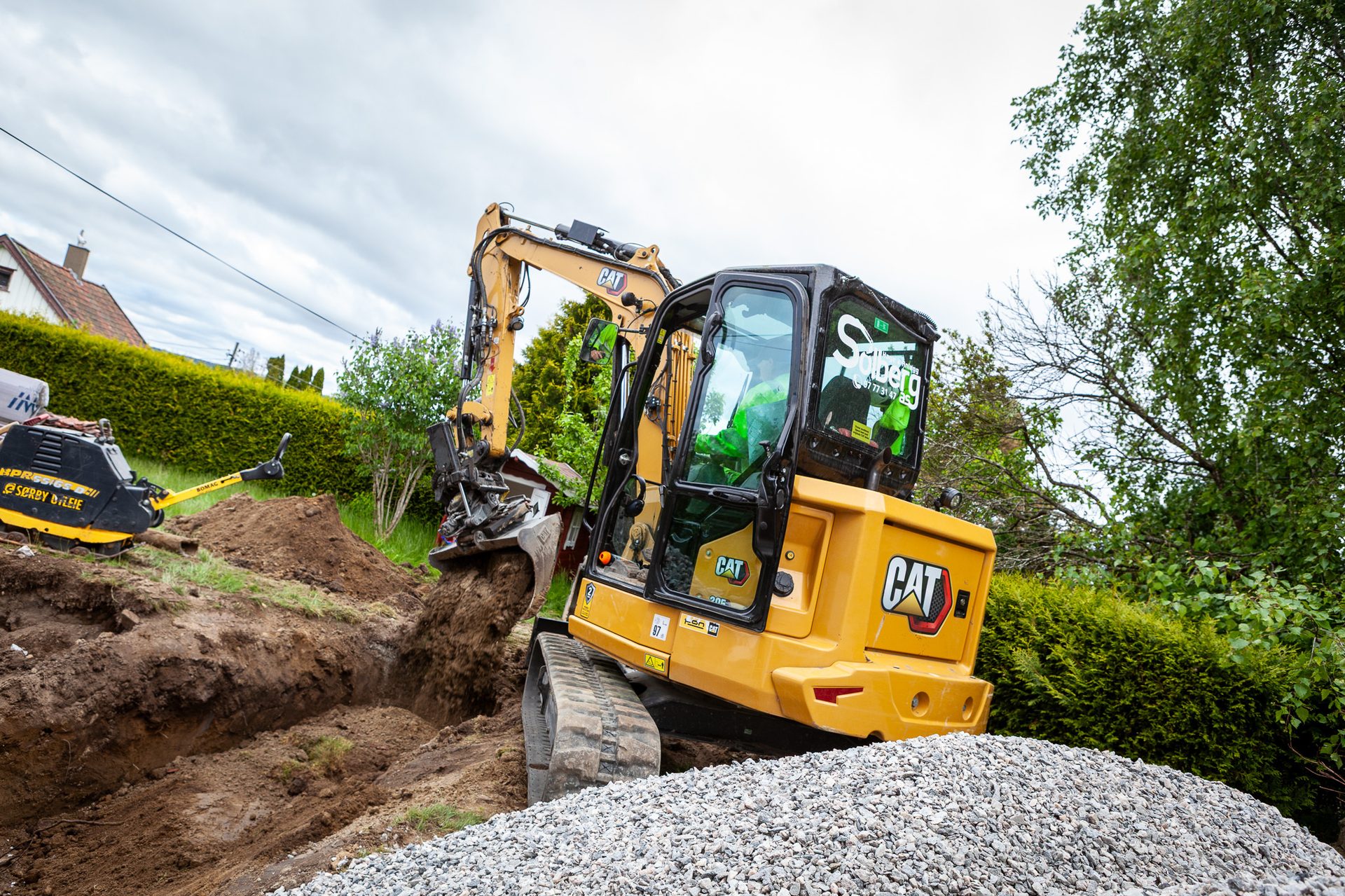 Yellow CAT excavator digging a trench, with a gravel pile and compactor.
