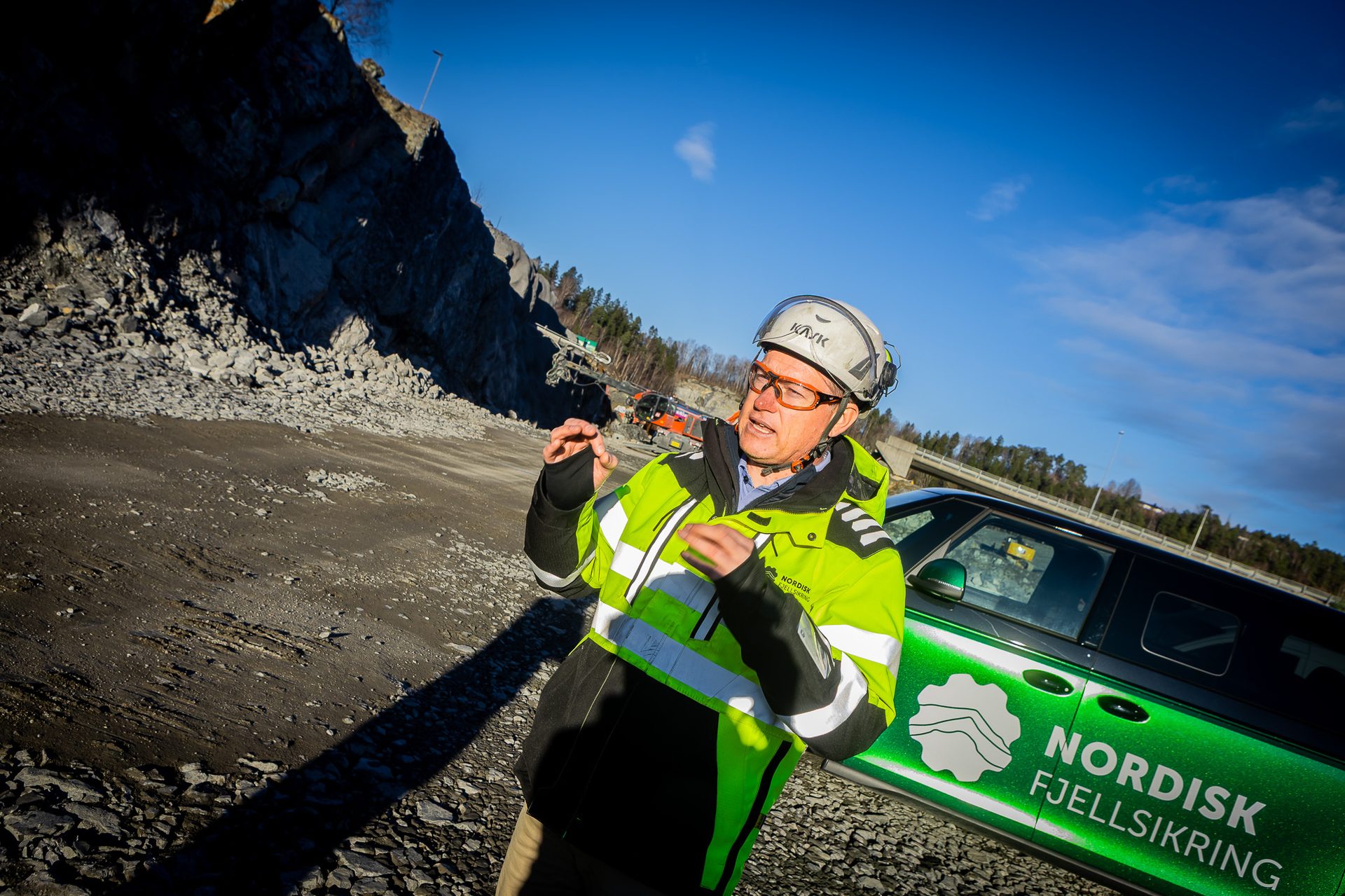 Man in high-vis vest, helmet, gesturing at rocky site with Nordisk Fjellsikring car.