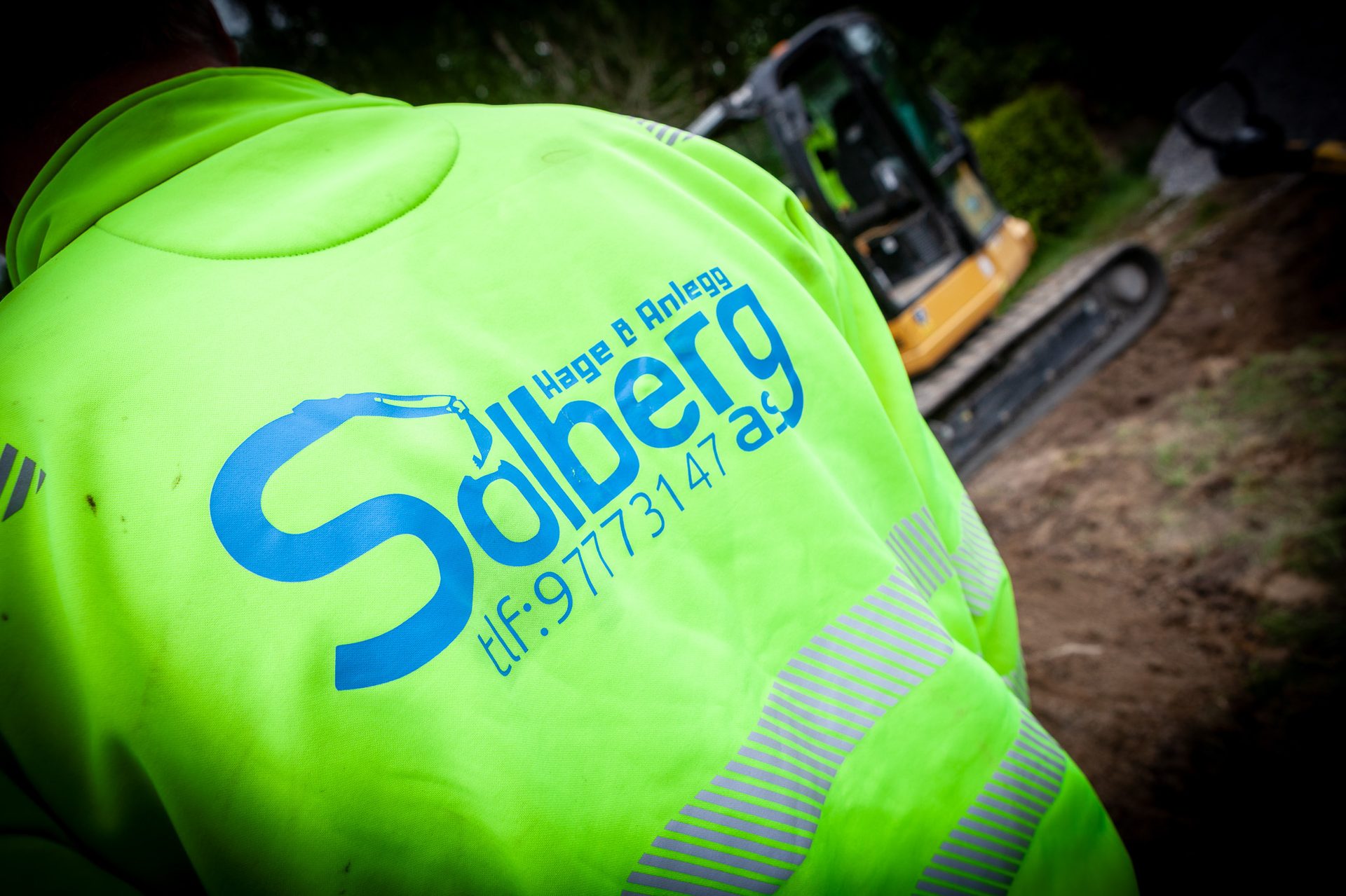 High-vis green jacket with 'Solberg' logo, worn by worker at construction site with excavator.