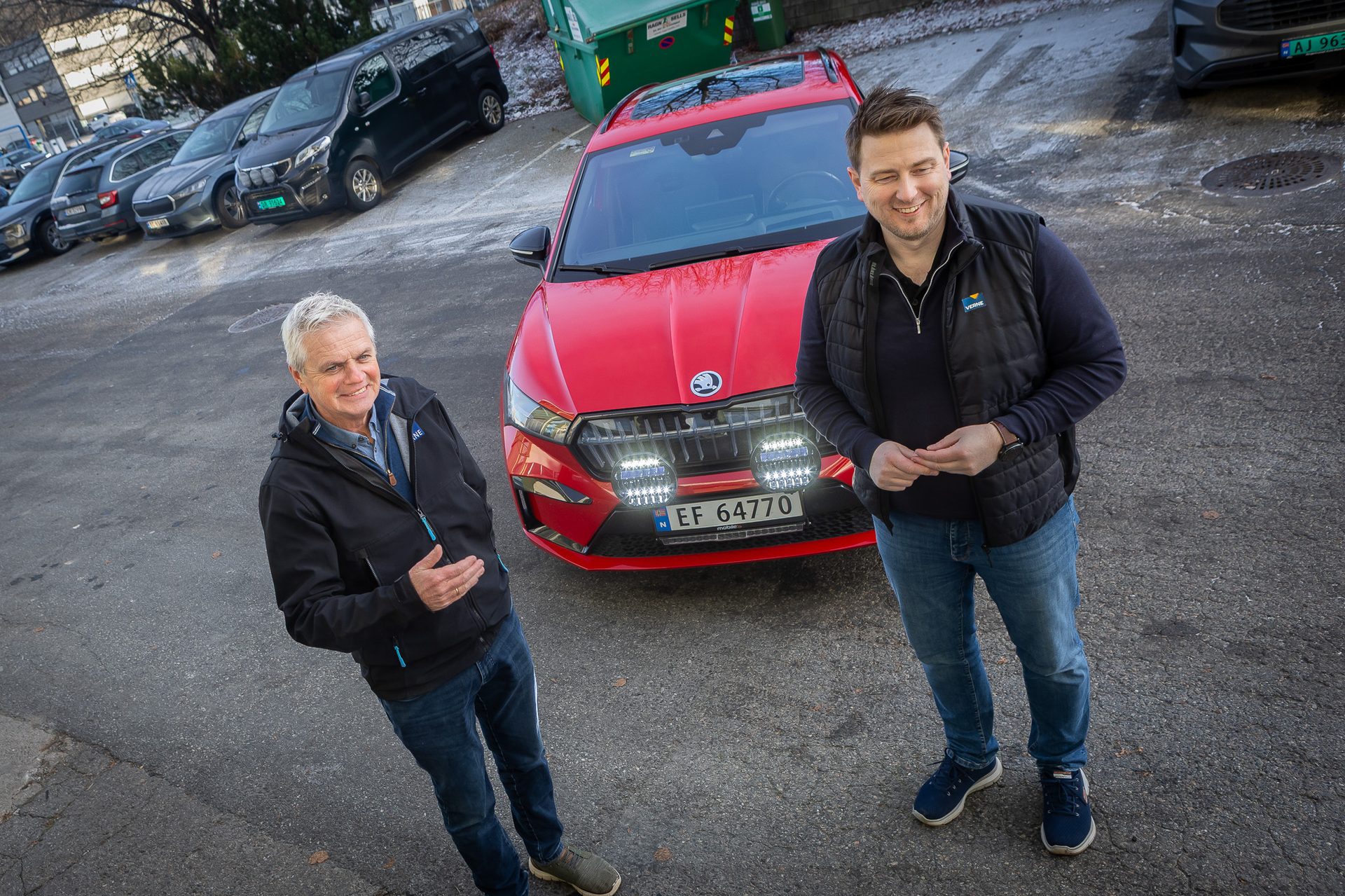 Two smiling men, one winking, stand by a red Skoda with bright rally lights.