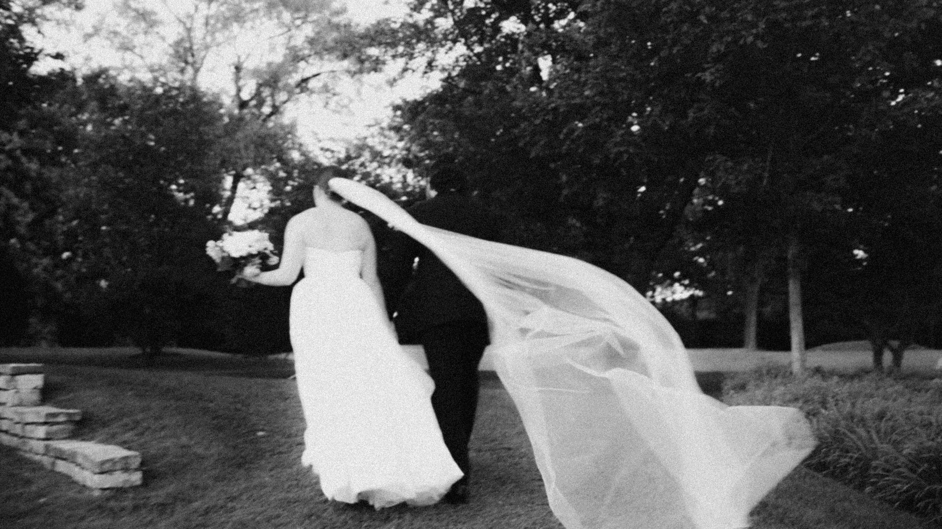 Black and white photo of a wedding couple walking away, the bride's long veil flowing in the wind.