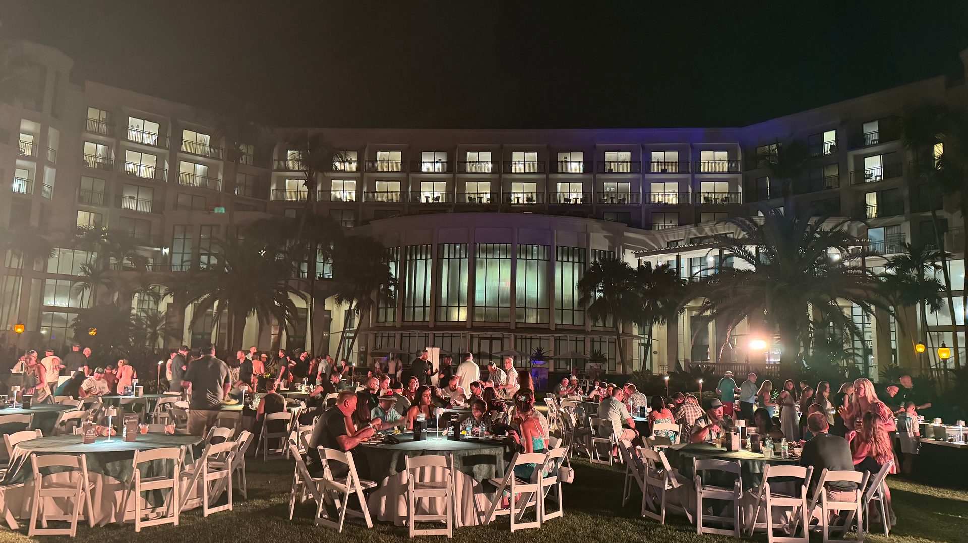 Outdoor night event: people at tables on a lawn, bright hotel & palm trees behind them.