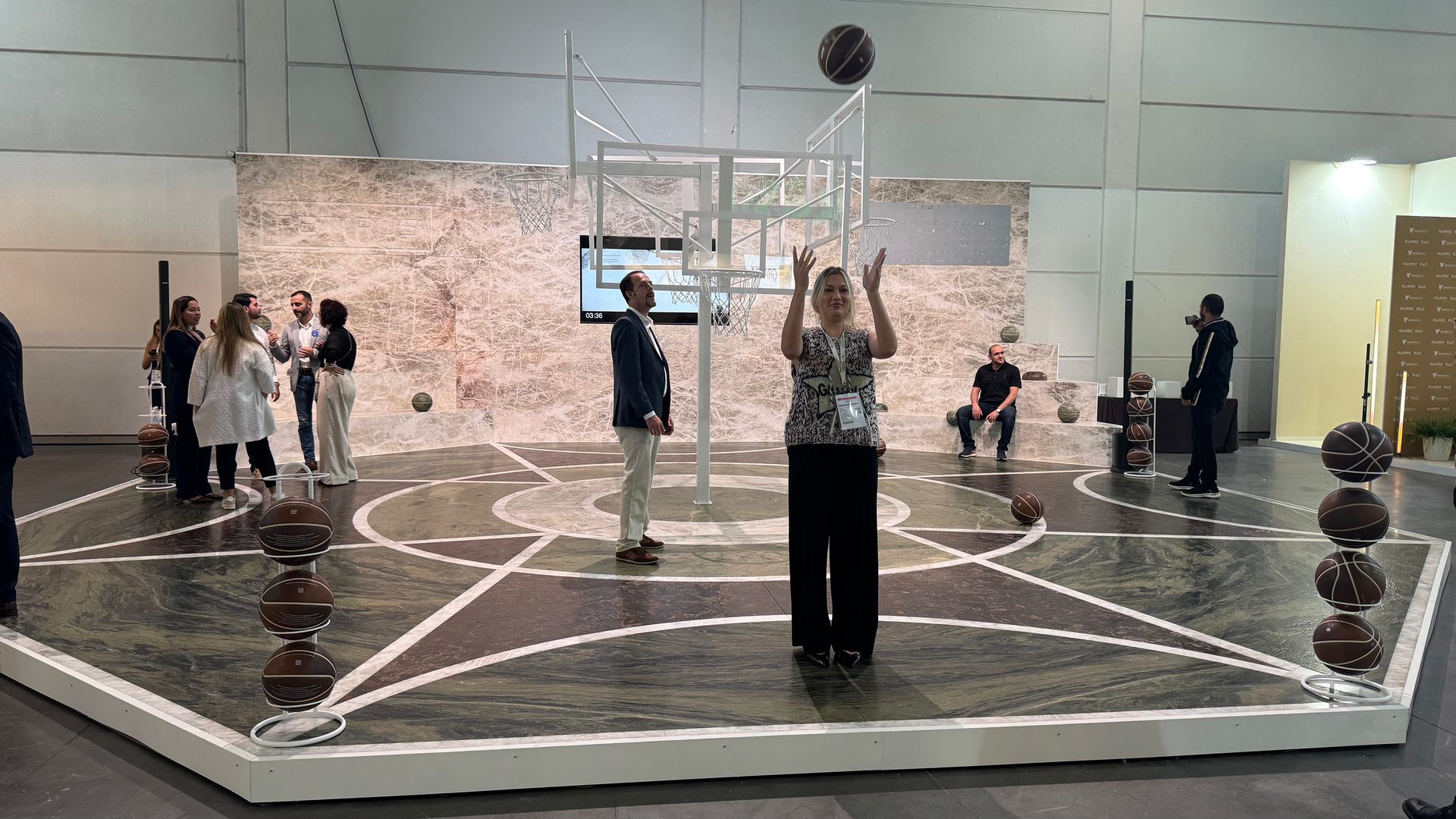 Woman throws basketball on a marble-patterned court with other people at an event.