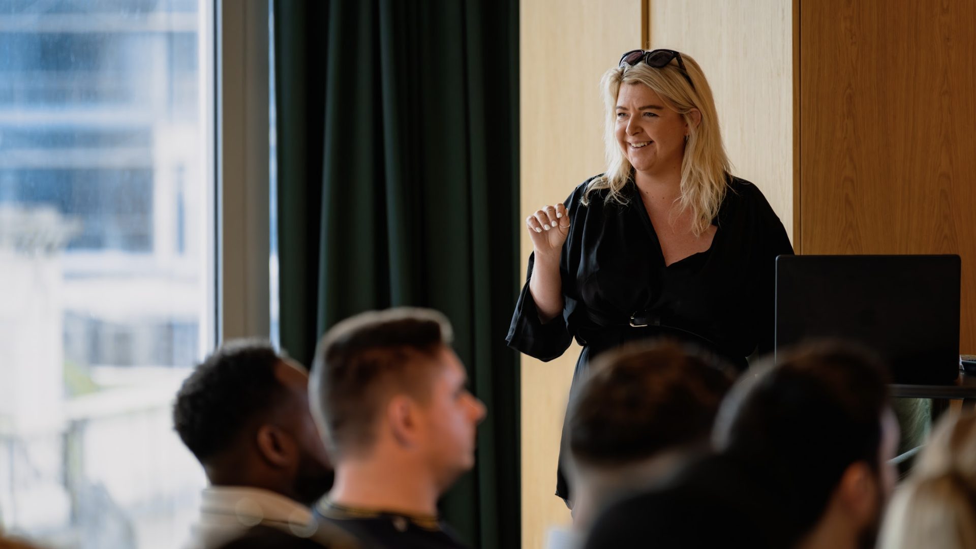 Blonde woman in black dress, smiling and gesturing, speaks to an audience; laptop next to her.