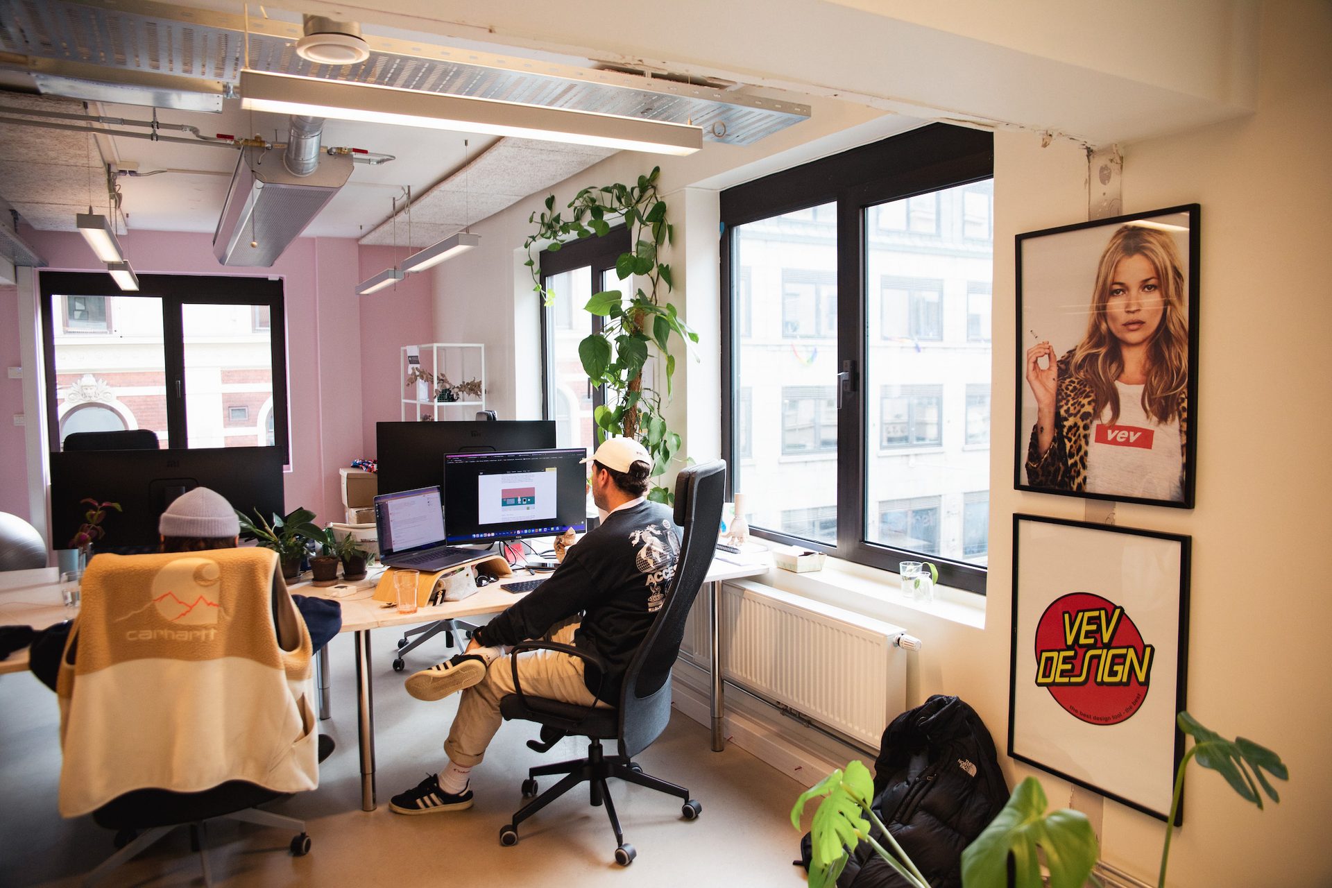Modern office with two people working at computers, featuring plants, large windows, and wall art.