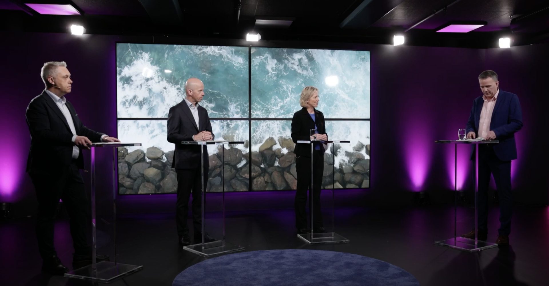 Four people stand at podiums on a purple stage with a video wall displaying ocean waves.