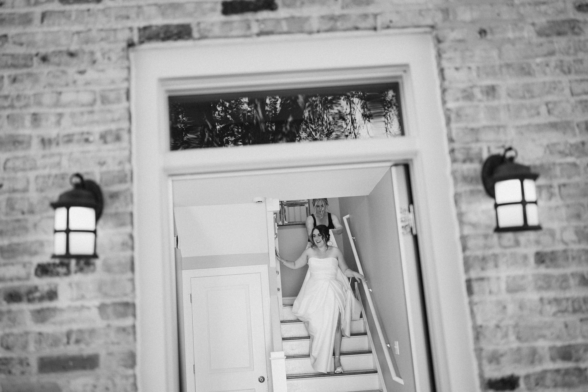 Black and white photo of a bride descending stairs, assisted by a woman, framed by a doorway.