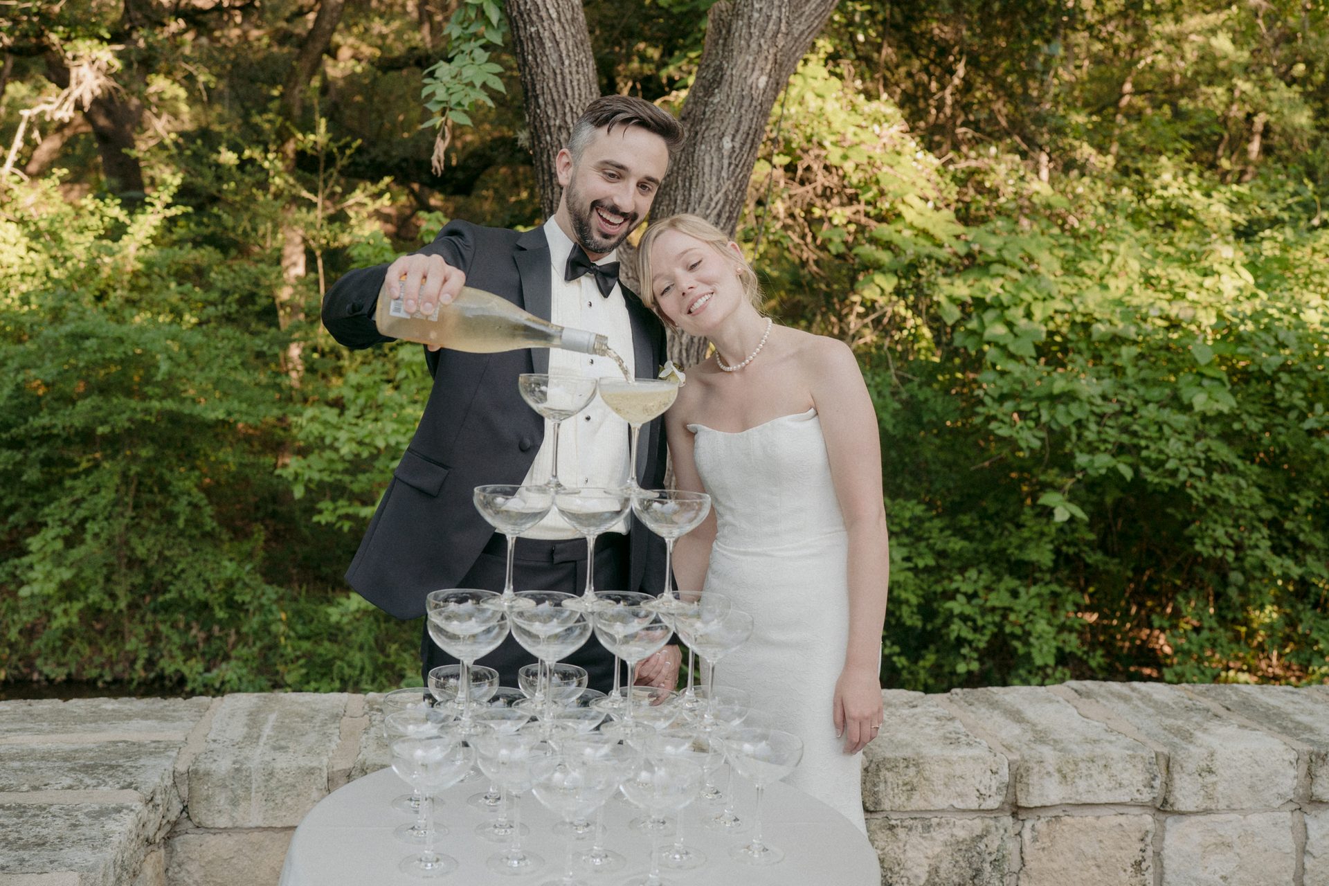 Groom pours champagne into a glass tower, bride smiles beside him, outdoors.
