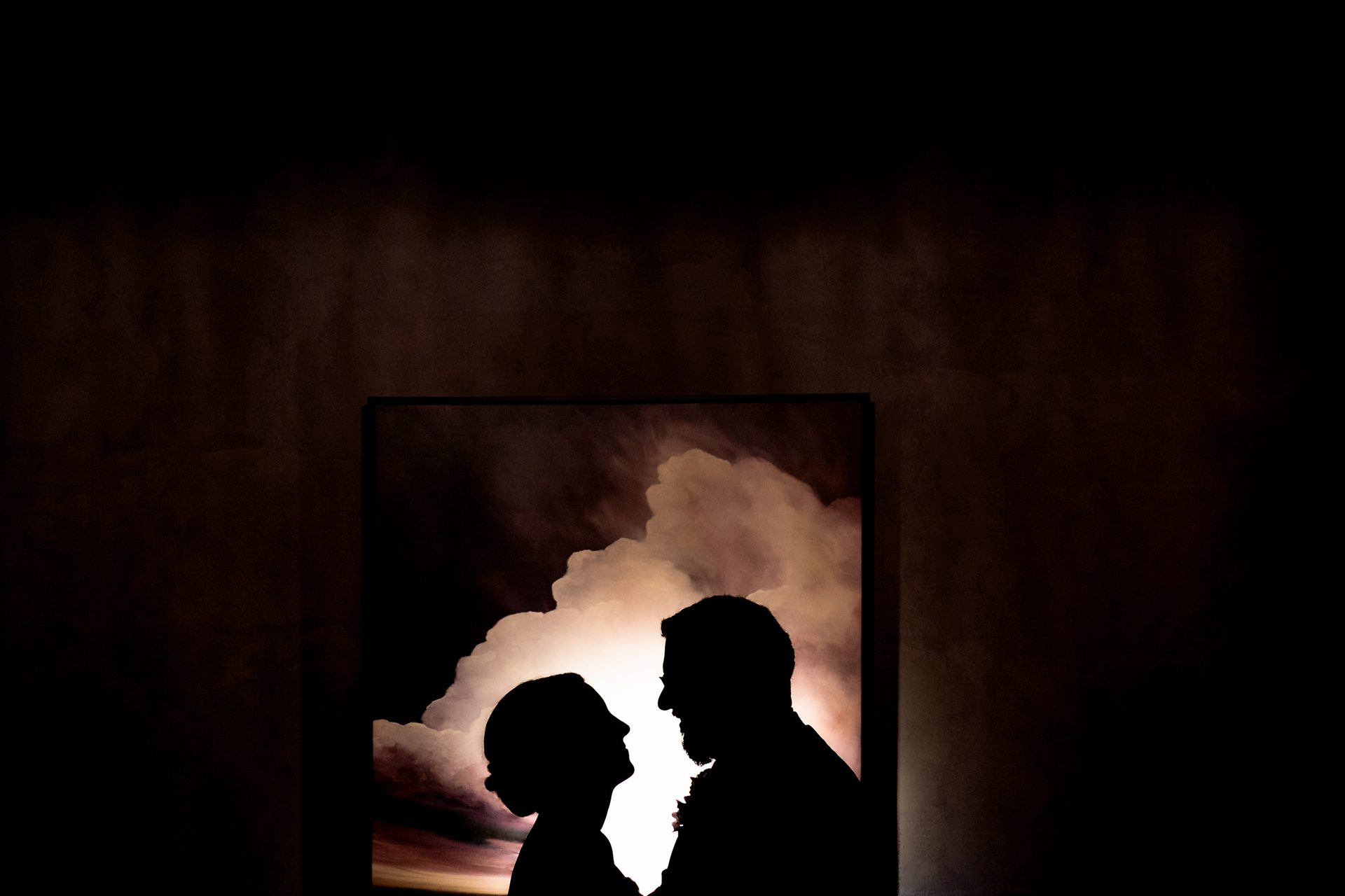 Silhouetted couple backlit by dramatic clouds within a dark, textured room.