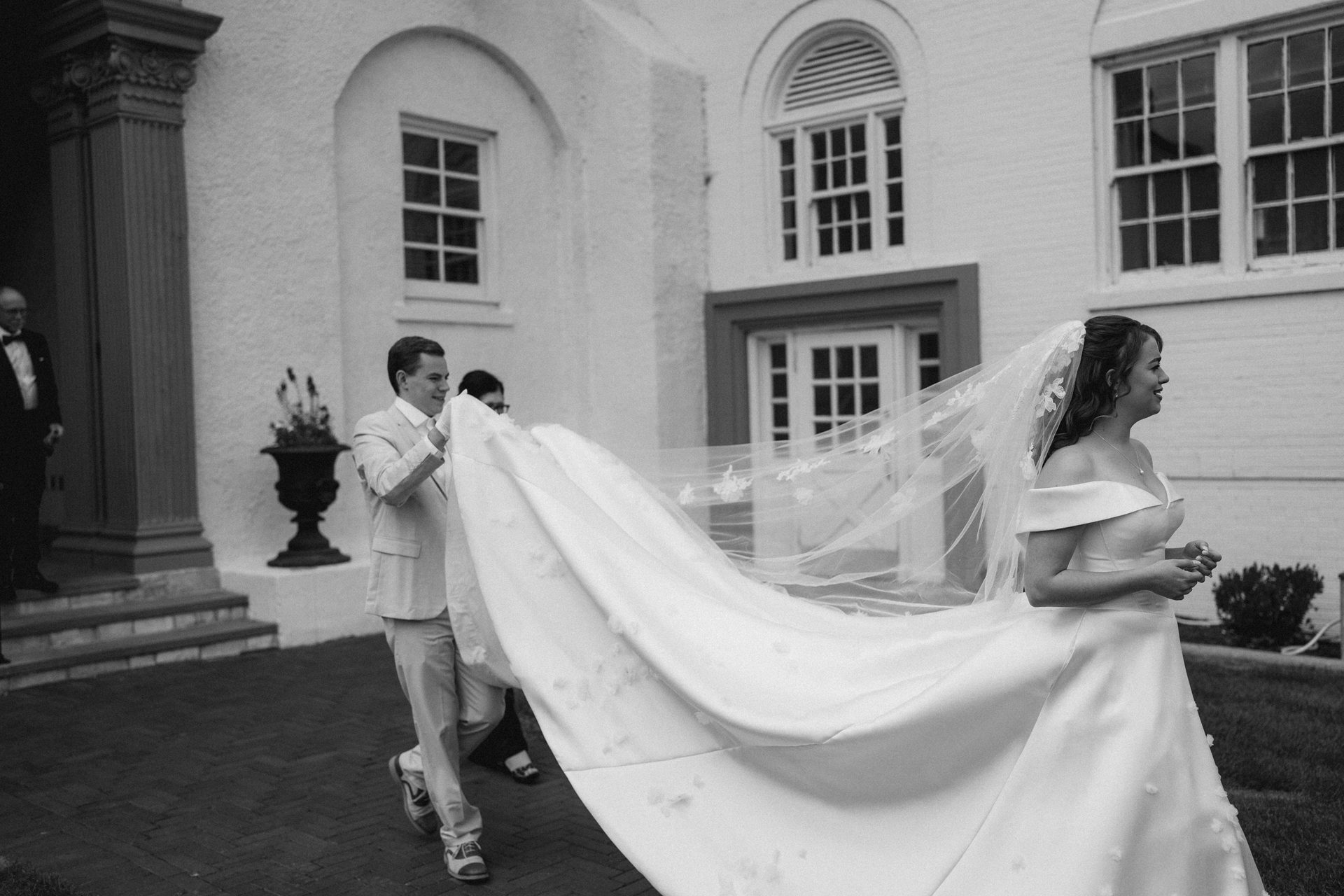 Black and white: Smiling bride in white dress, a man holds up her long train.