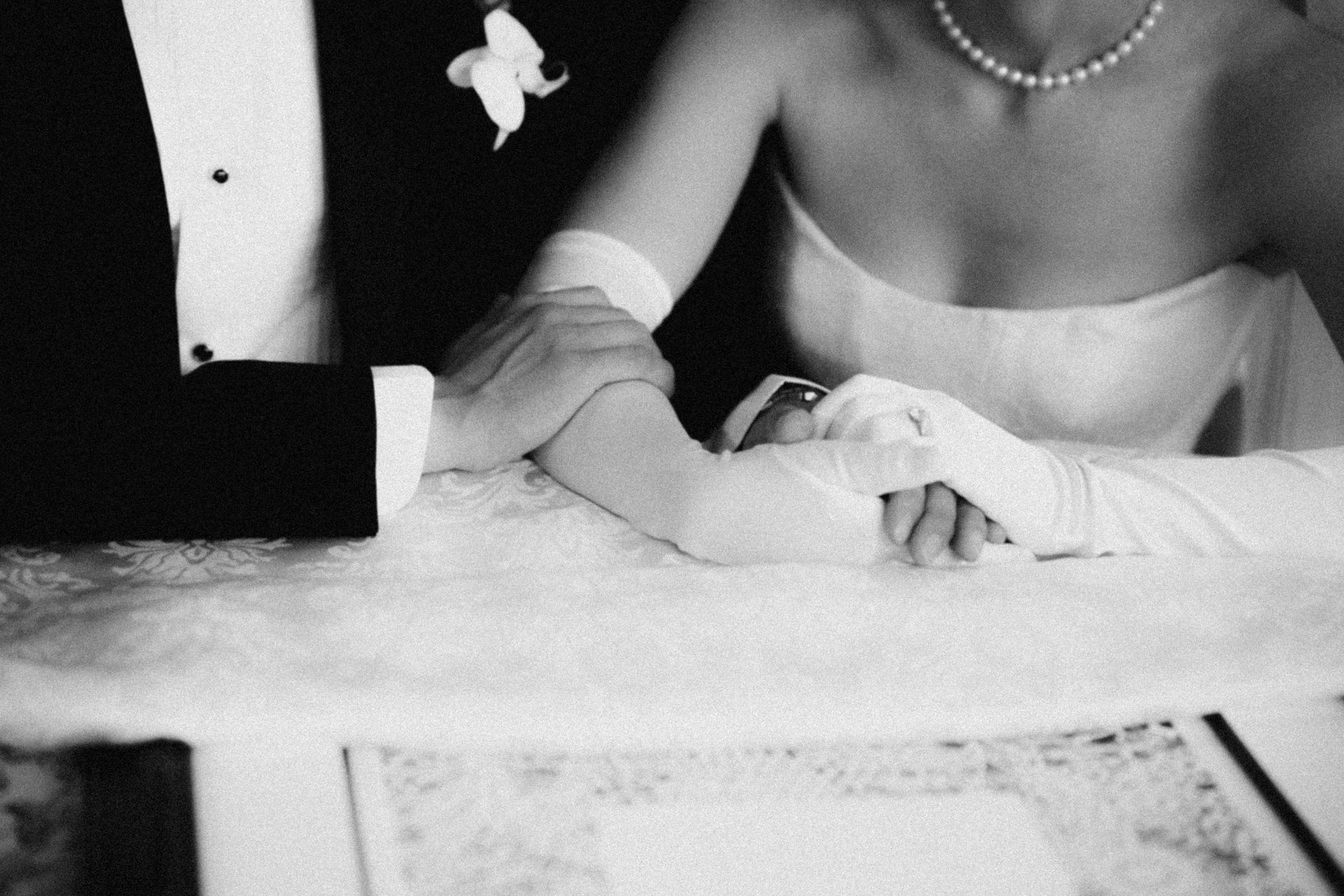 Black and white close-up of a bride and groom's hands clasped, showing a ring and glove.