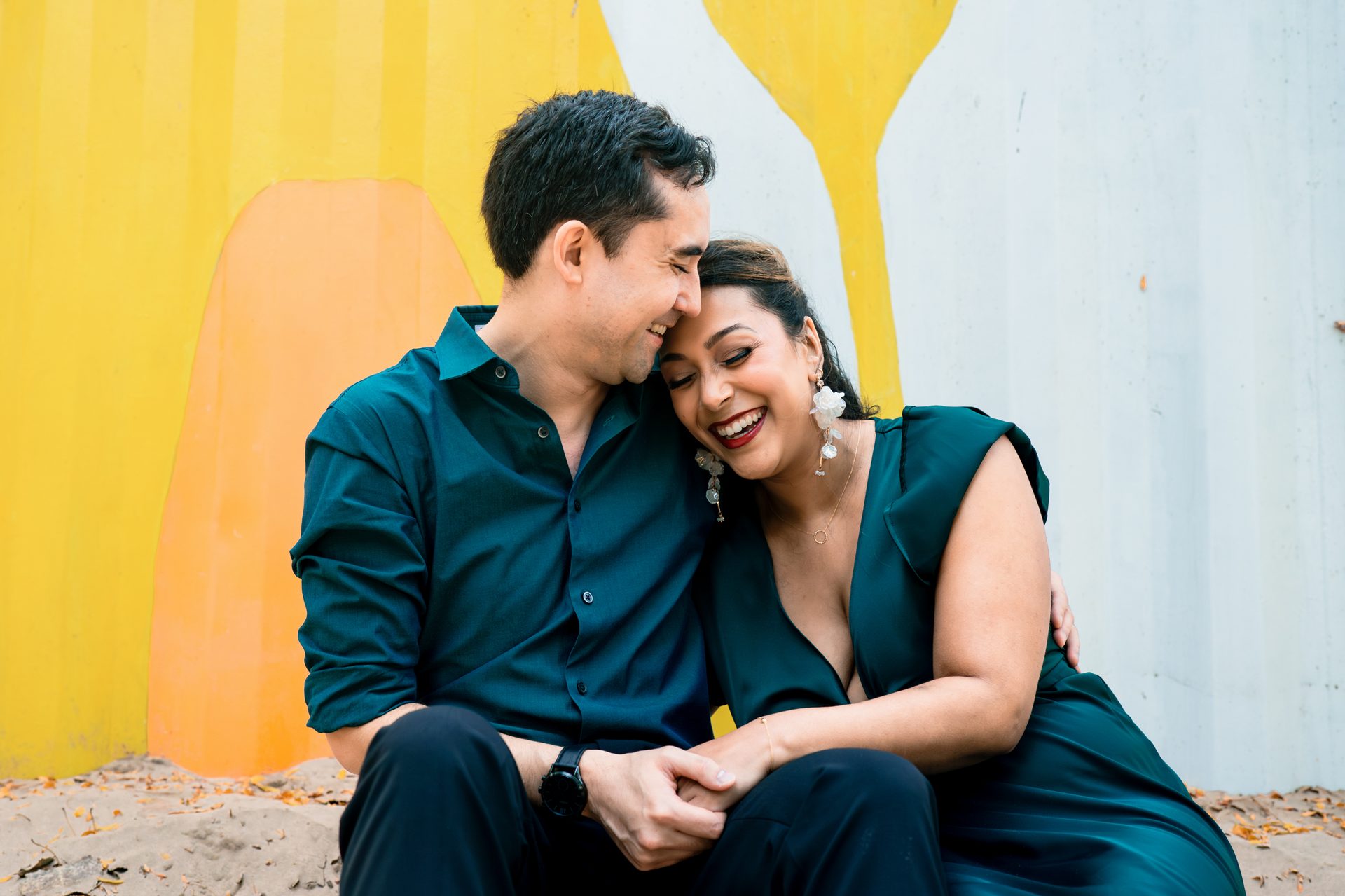 Joyful couple in dark green embrace, laughing against a colorful wall.
