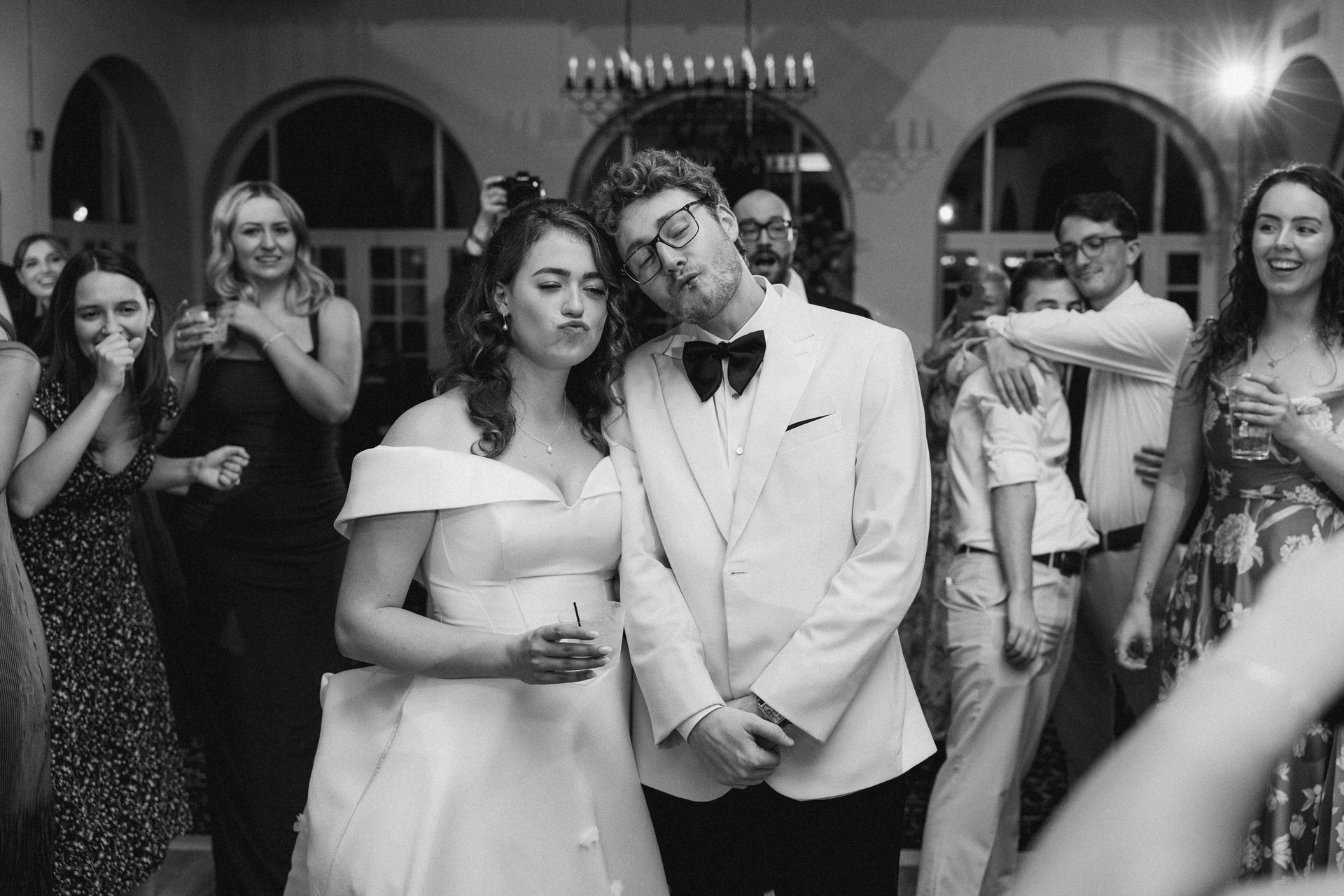 Black and white photo: a bride and groom make silly faces, surrounded by guests.