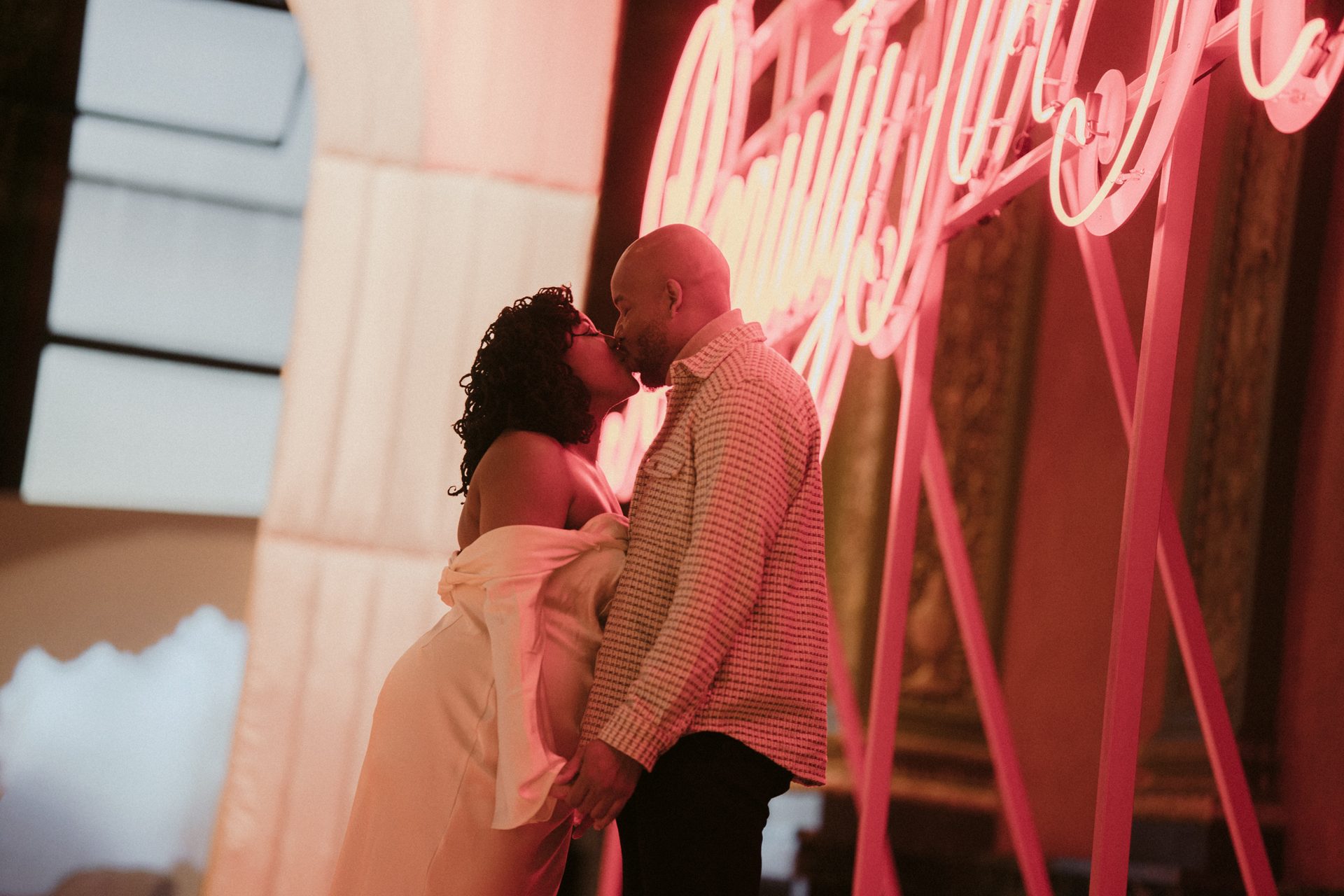 A couple kisses under a pink neon sign, bathed in its glowing light.