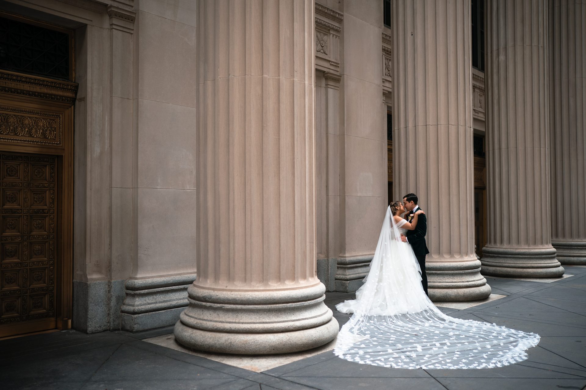 Bride and groom kiss between large columns, long wedding dress train spread out.