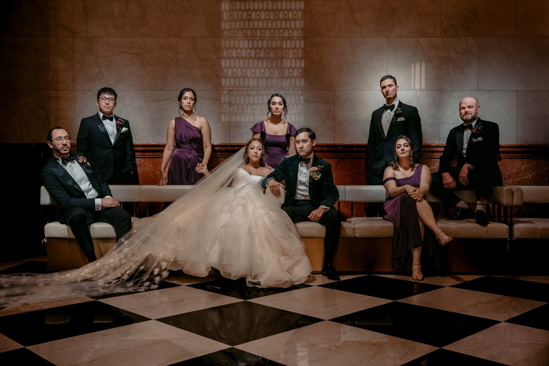 Formal wedding party portrait with bride, groom, and six guests on a checkered floor.