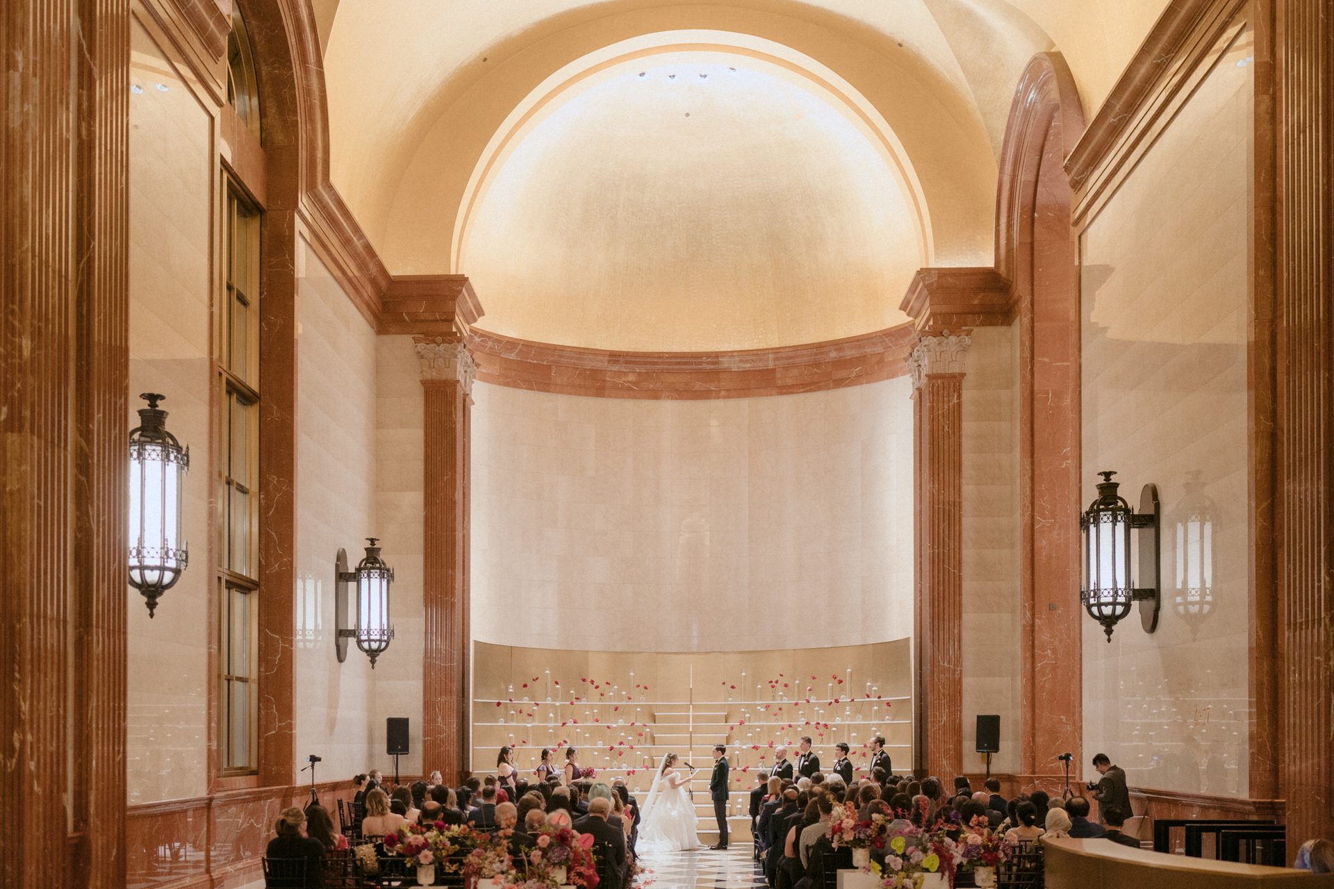 A wedding ceremony in a grand hall with high arches and columns, featuring the bride, groom, and guests.