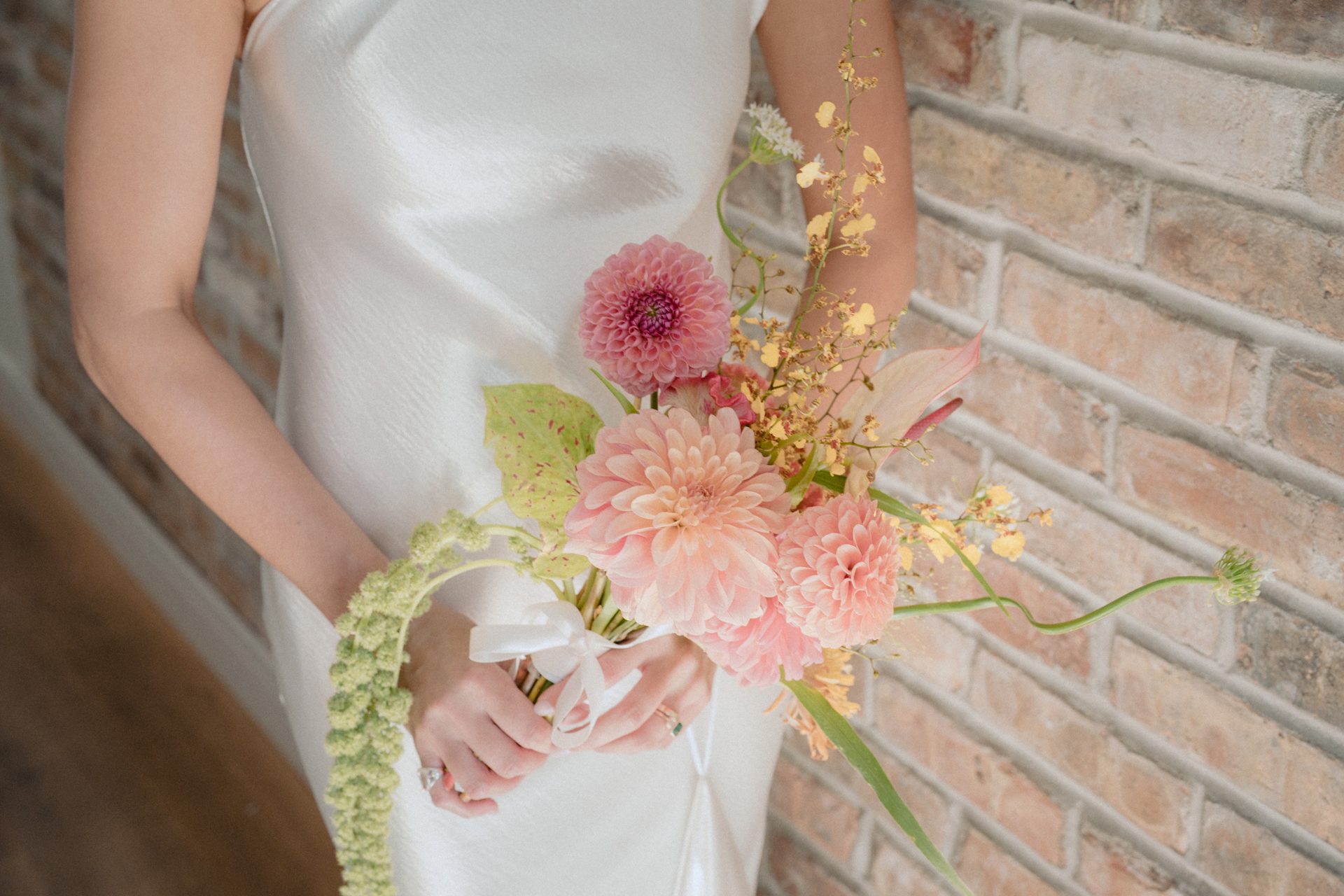 Hands in a white dress hold a vibrant pink and peach flower bouquet against a brick wall.