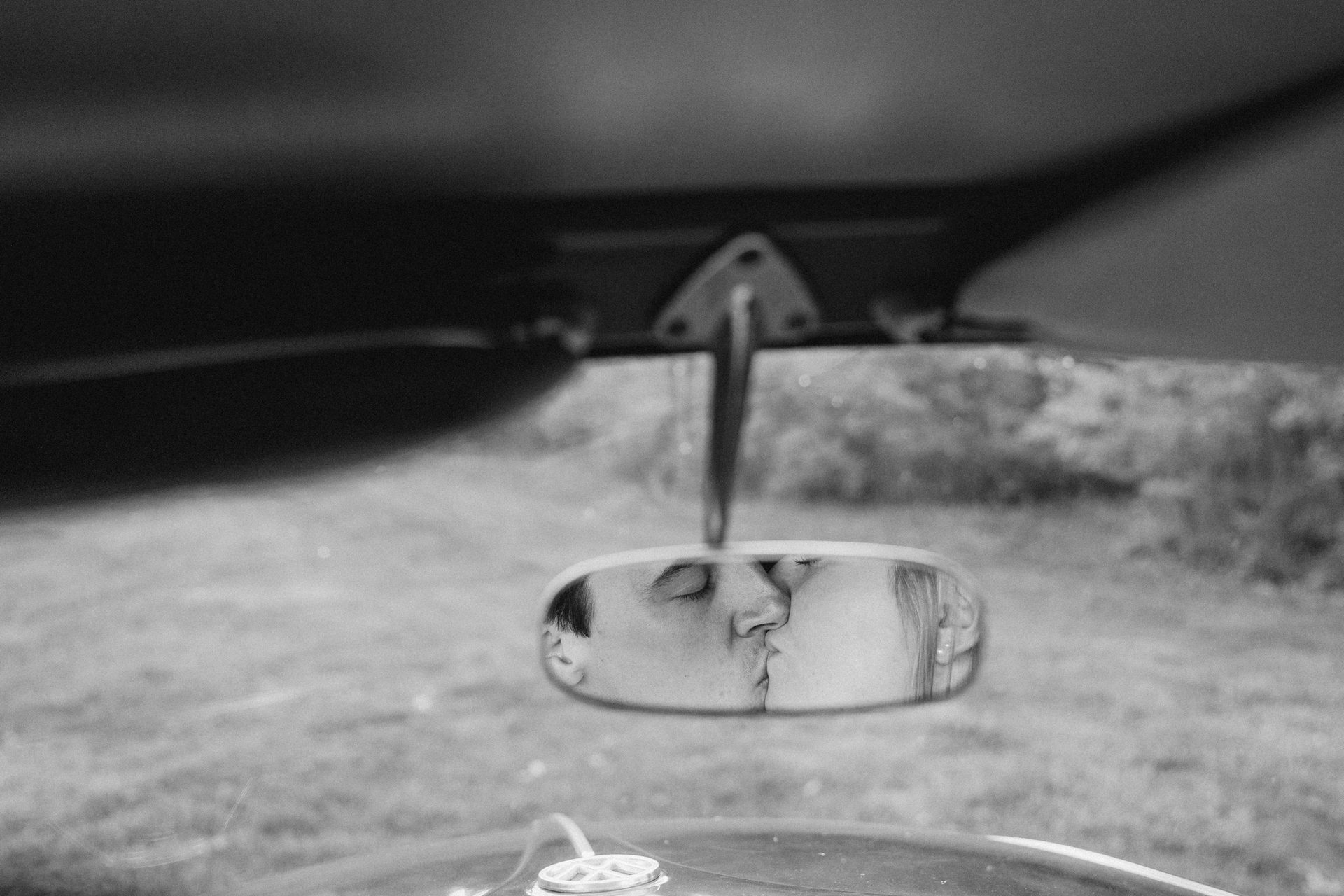 Black and white photo of a couple kissing in a car's rearview mirror.