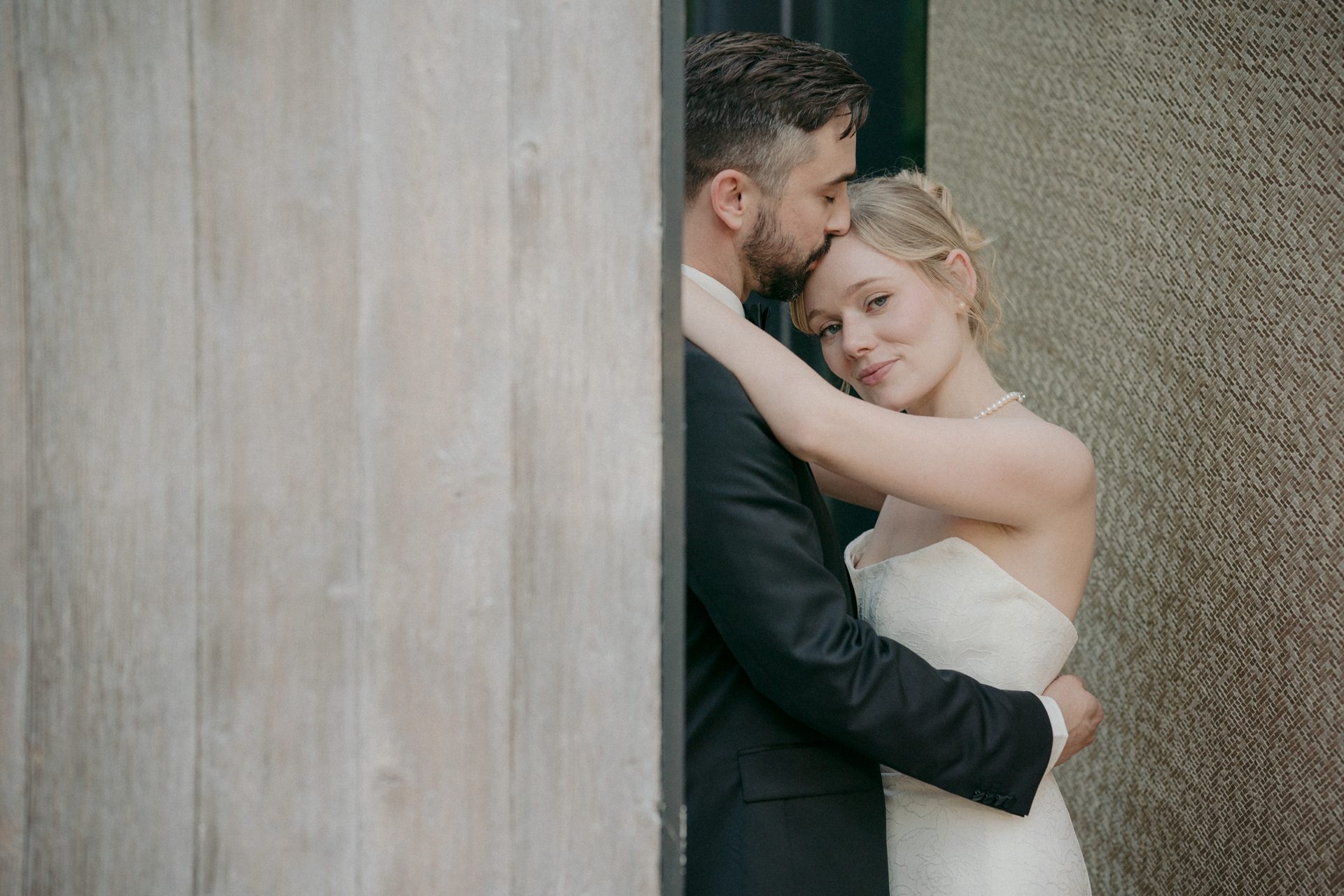 Bride and groom embracing. He kisses her forehead, she looks at the camera.