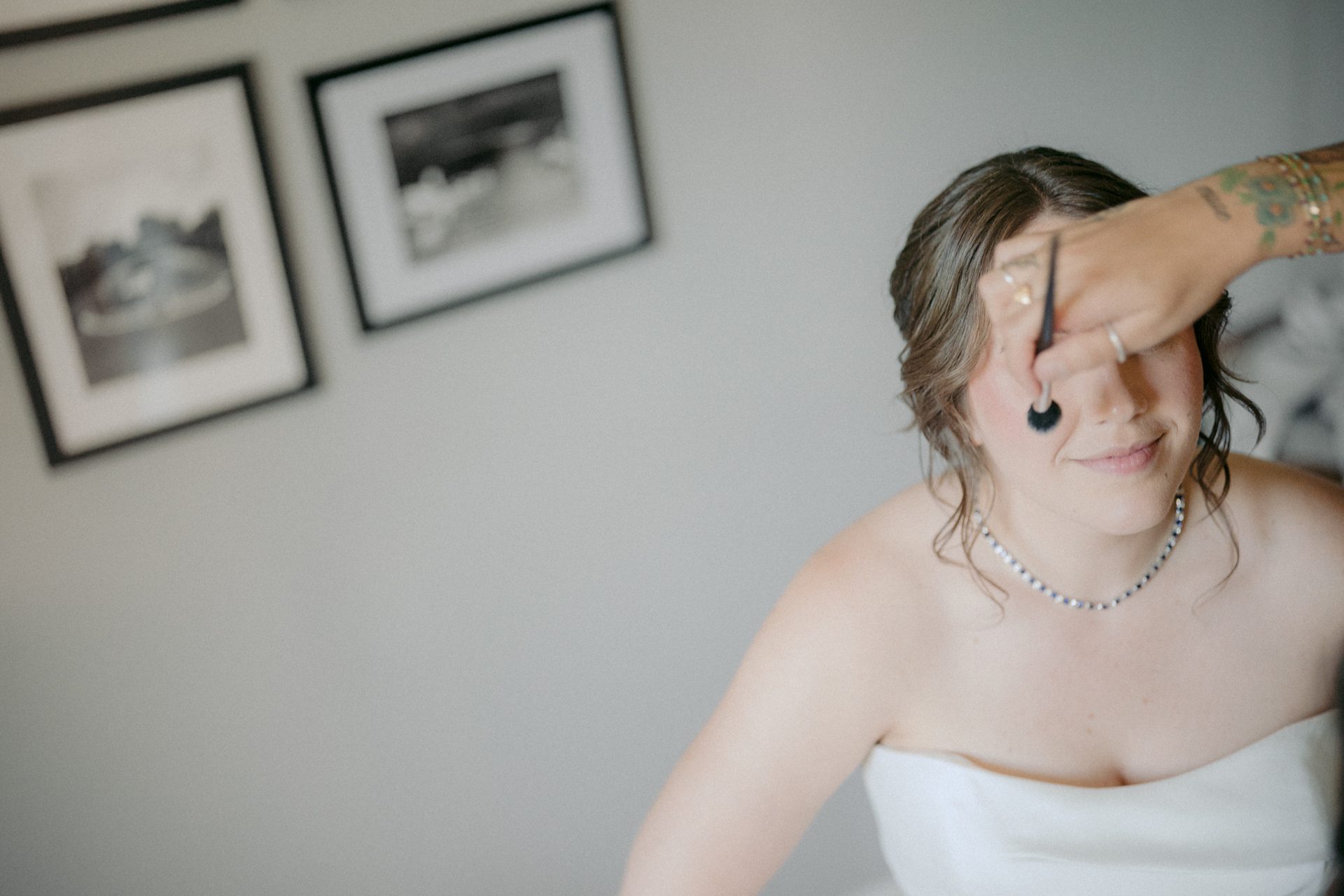 Bride smiling as makeup is applied with a brush, wearing a white dress and necklace.