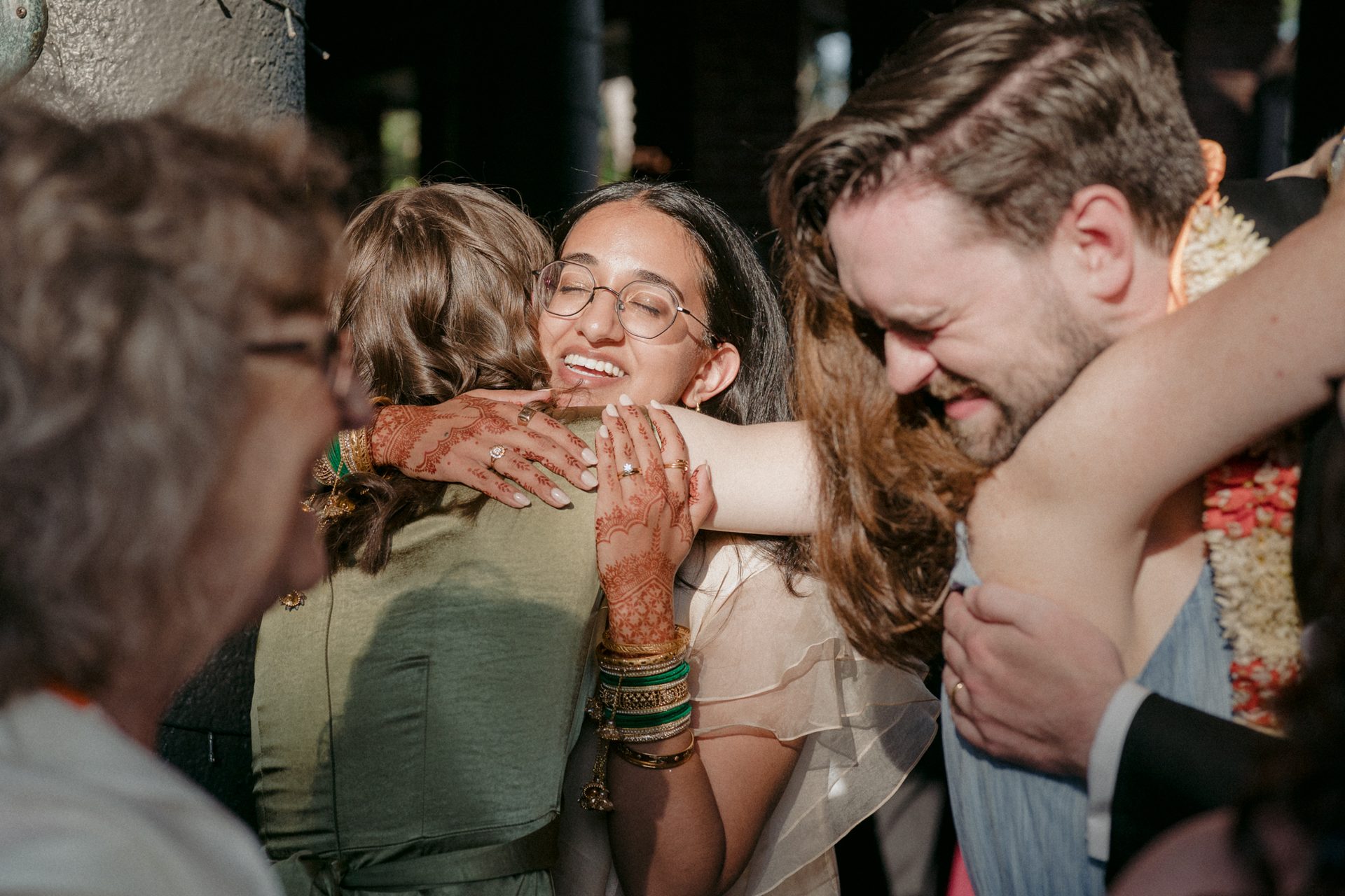 Joyful woman with henna hands embraces two people; a man also hugs others.