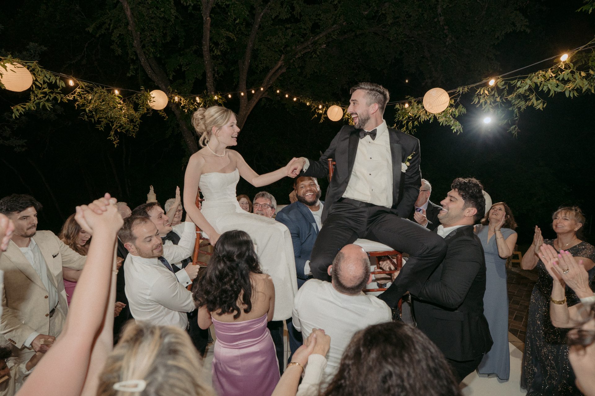 Joyous bride and groom lifted on chairs, holding hands, smiling at their night wedding with cheering guests.