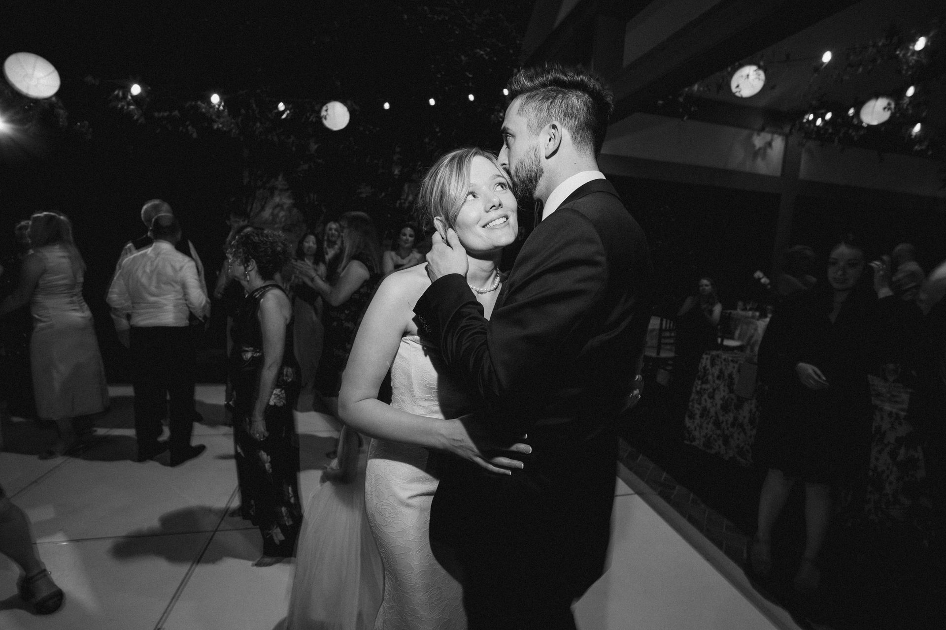 B&W photo: Affectionate couple slow dancing at night, bathed in string light glow.