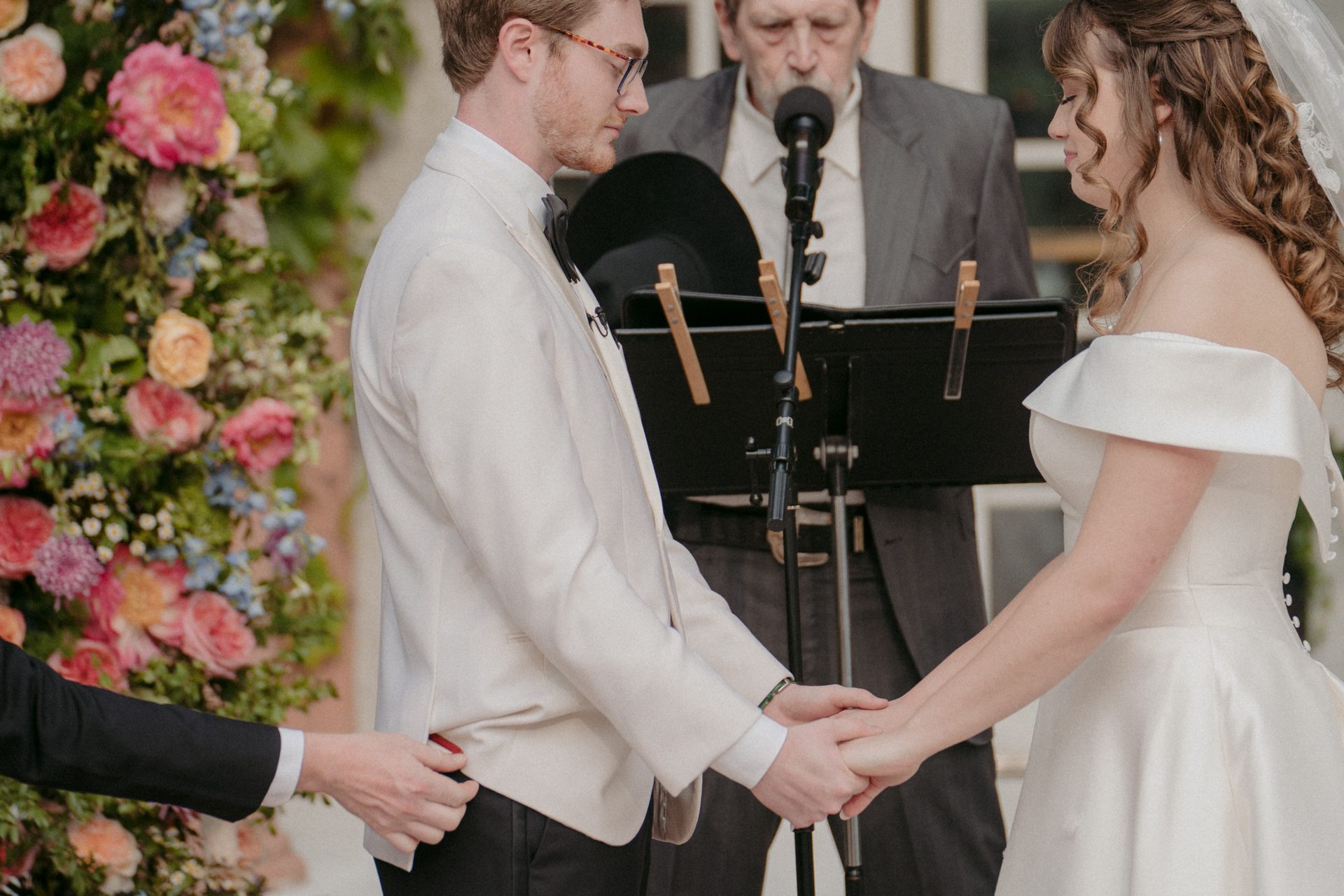 Bride and groom holding hands at their wedding ceremony, minister in background, another hand on groom's back.