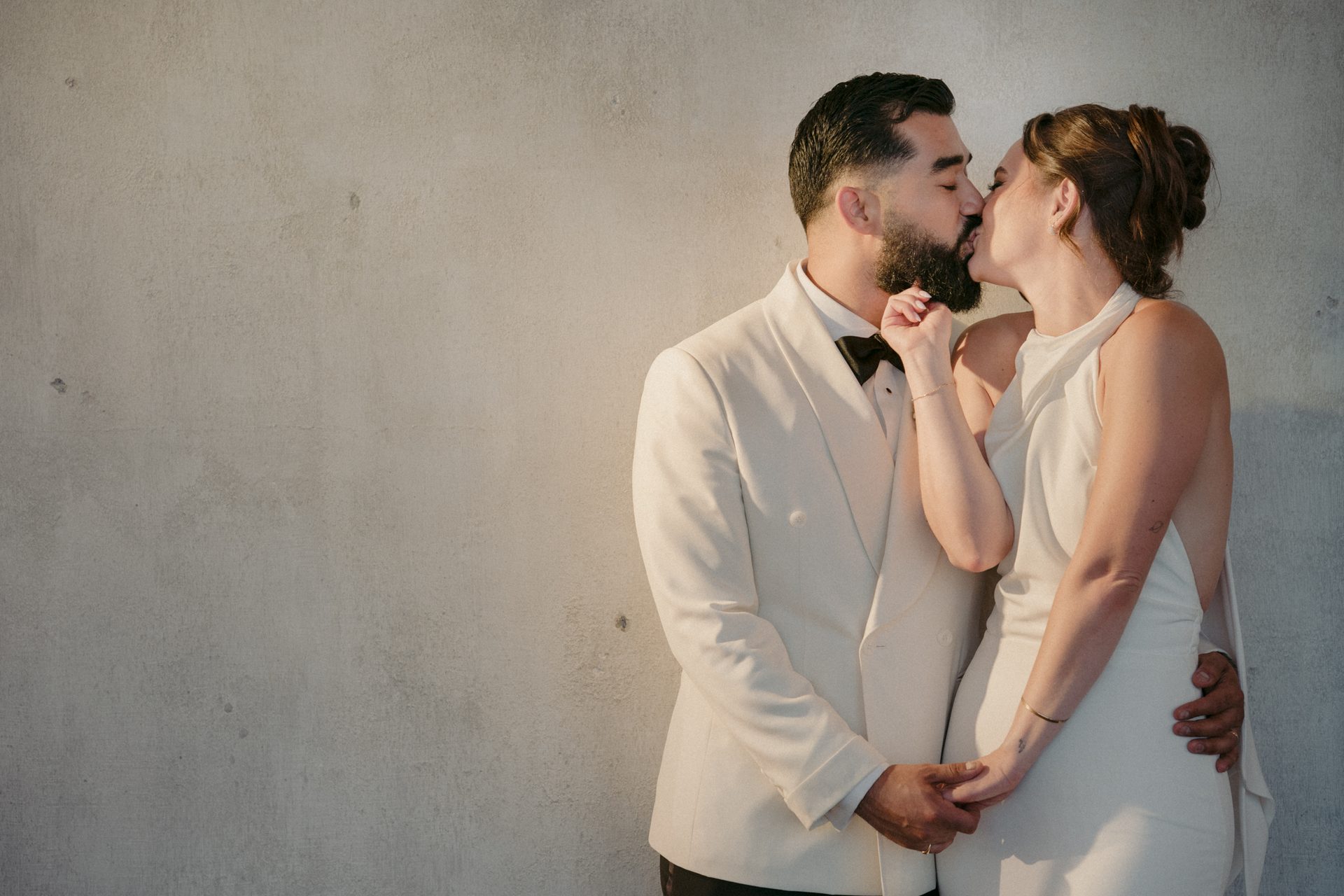 Wedding couple, man in white tuxedo, woman in white dress, kissing and holding hands by a concrete wall.