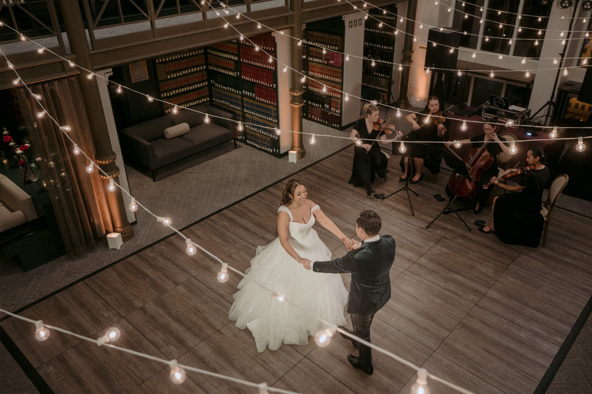 A couple dances under string lights, with a string quartet playing in a room with bookshelves.
