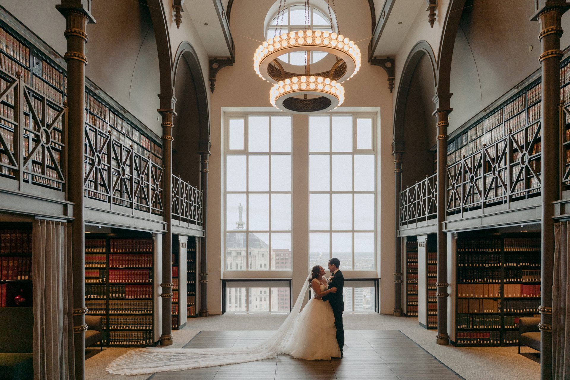 Bride and groom embrace in a grand library with two-story bookshelves, large windows, and chandeliers.