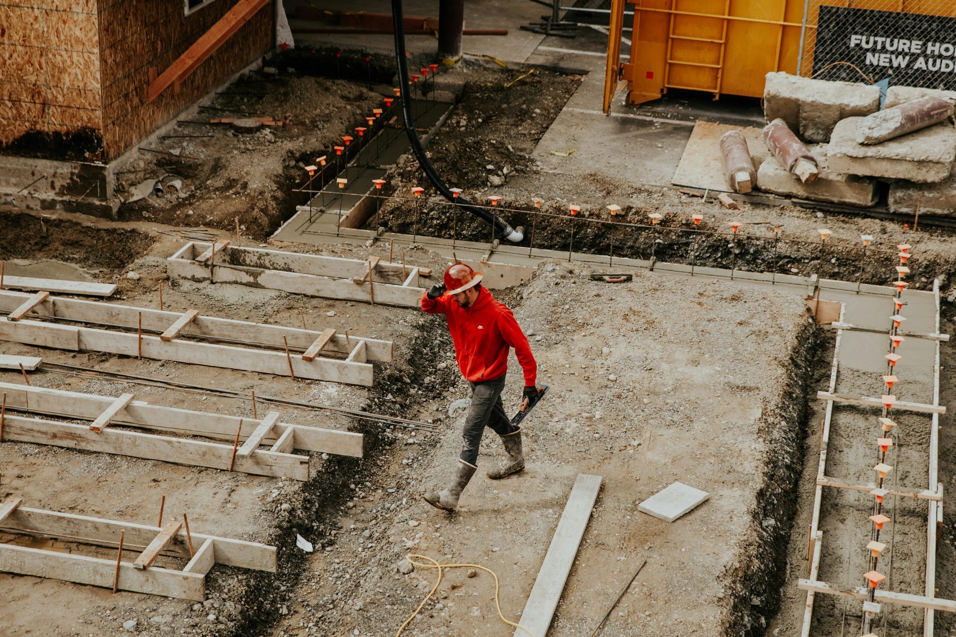 Man in a red hoodie and hard hat walks on a construction site with new concrete foundations.