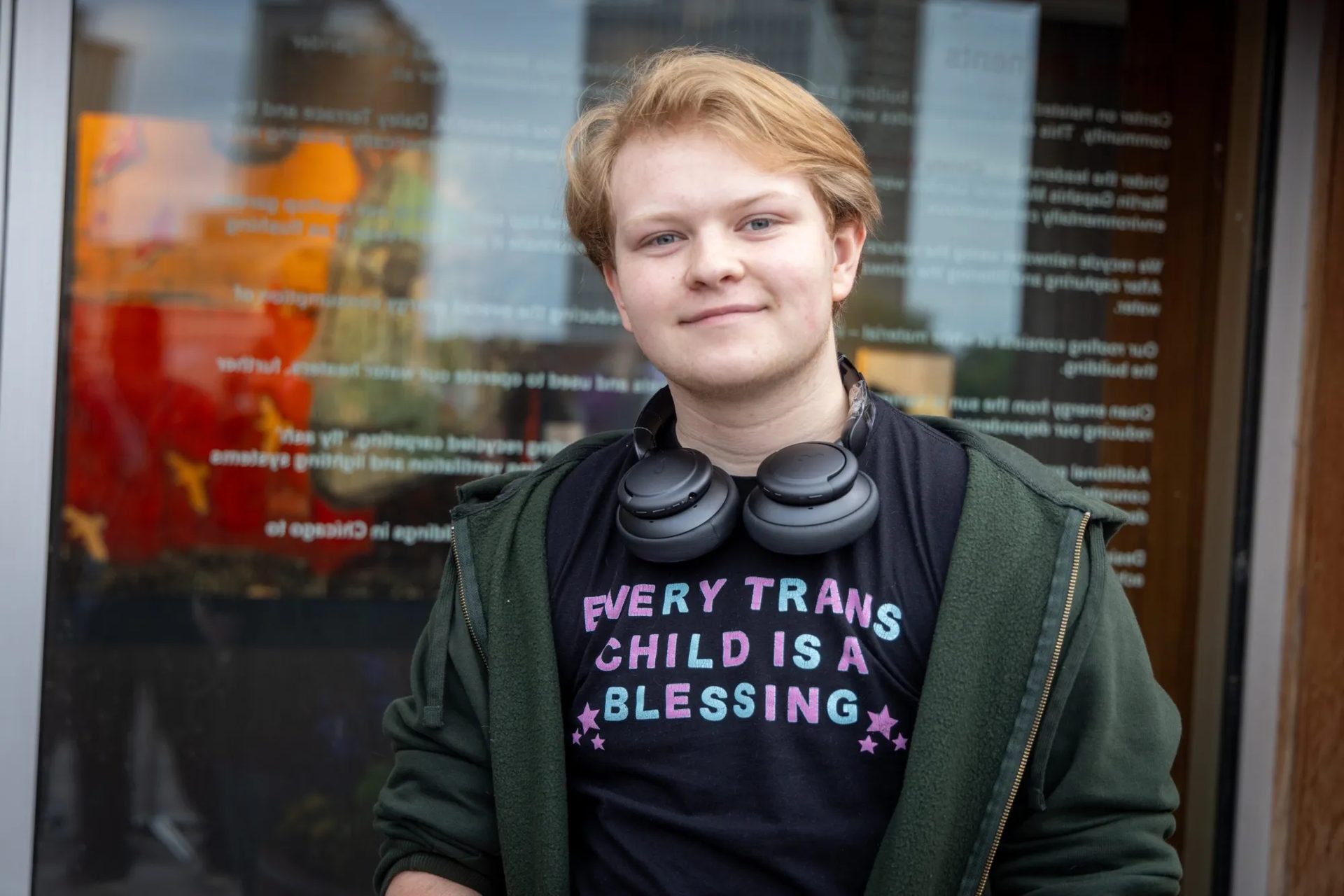 Smiling person in a "EVERY TRANS CHILD IS A BLESSING" t-shirt, headphones around neck.