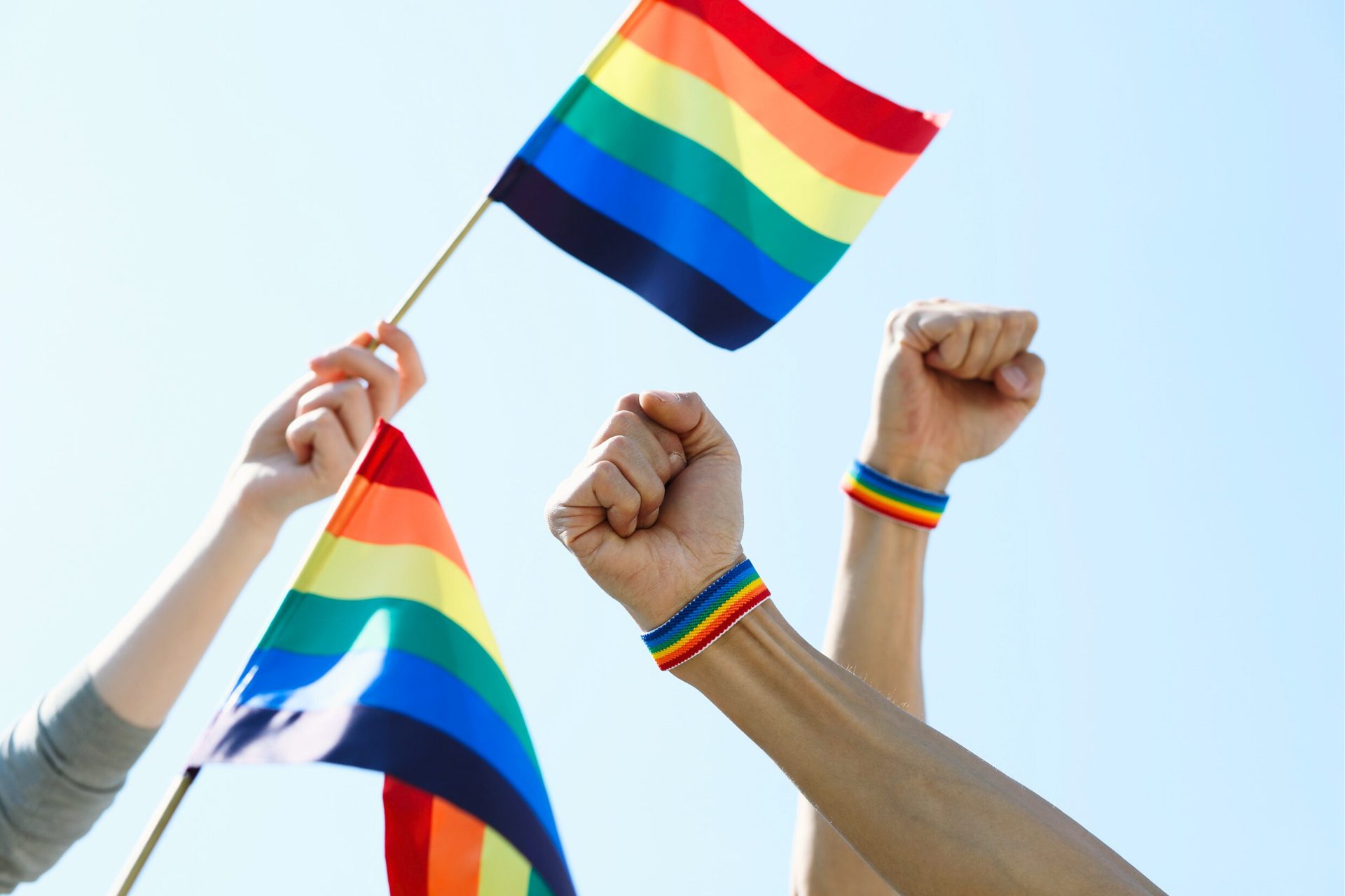 Raised fists with rainbow wristbands and hands holding rainbow flags, symbolizing LGBTQ+ pride.