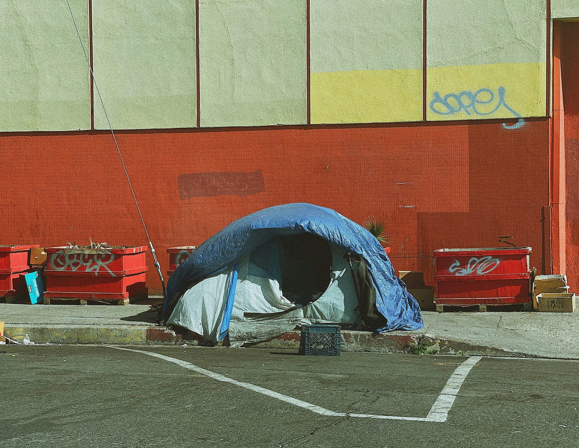 Blue tarp tent on a city sidewalk next to red planters and a brick wall with graffiti.