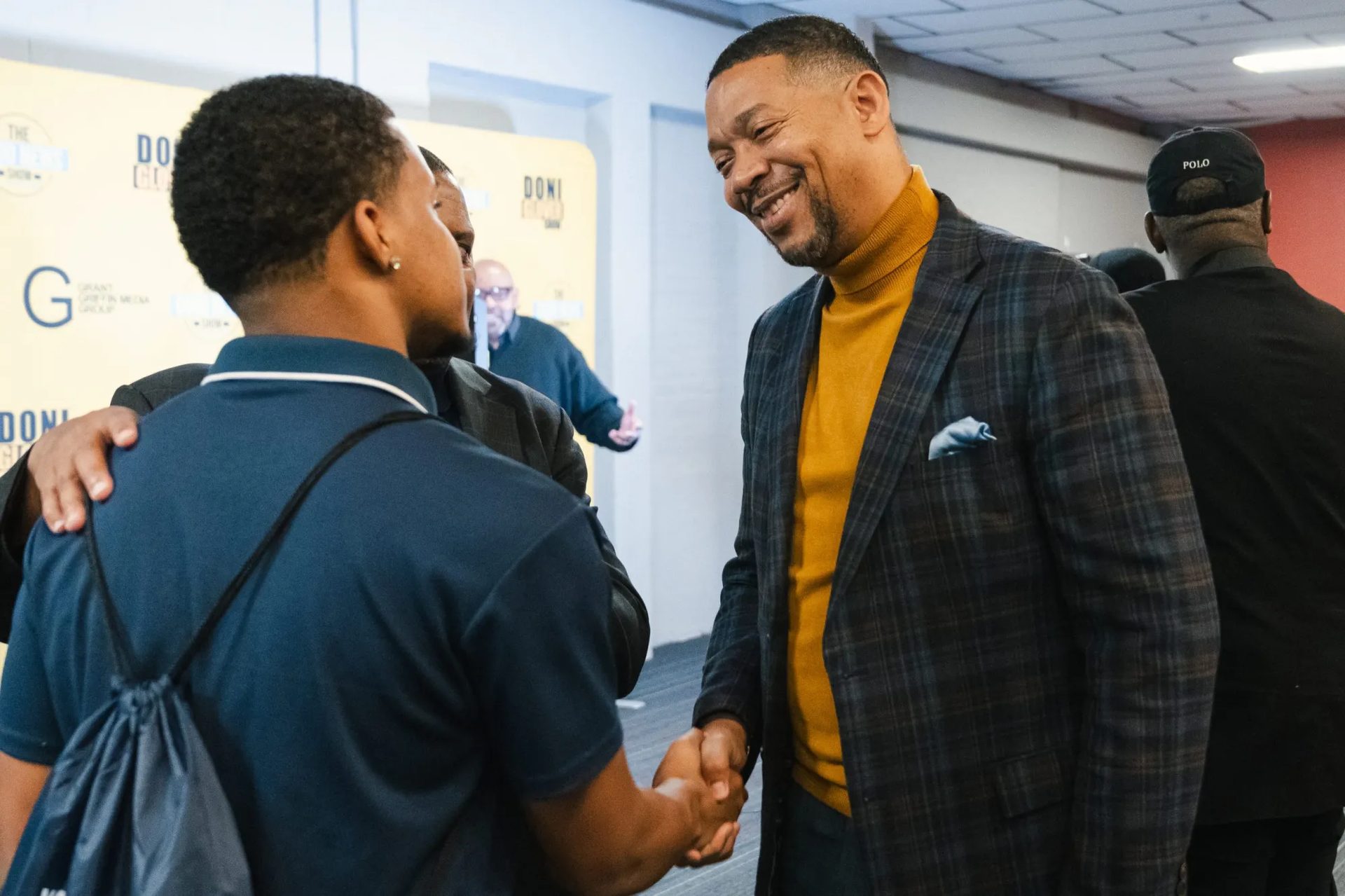 Two men, one smiling, shake hands in front of a branded backdrop.