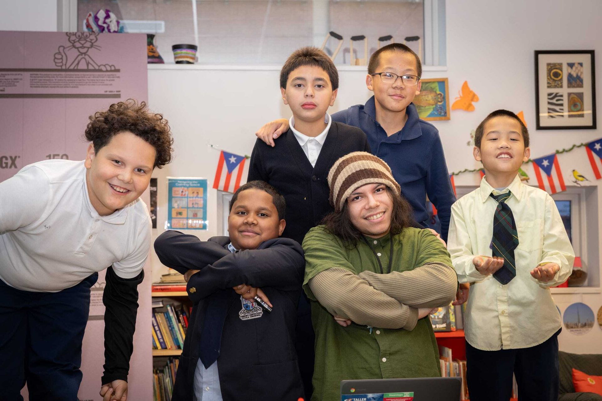 Six smiling children of diverse backgrounds pose together in a classroom setting, surrounded by flags and books.