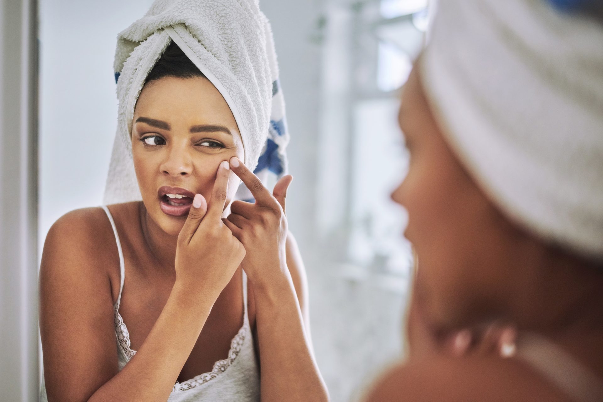 Woman in towel examining her face in mirror, touching eye area.