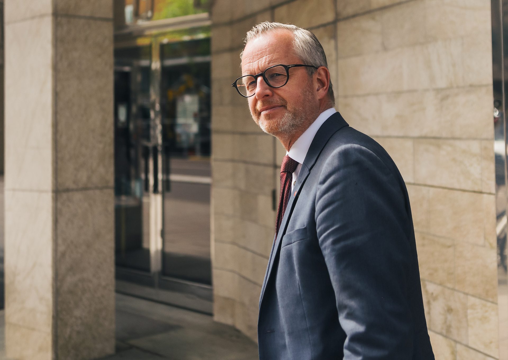 A smiling man in a suit and glasses with grey hair and beard standing outdoors.
