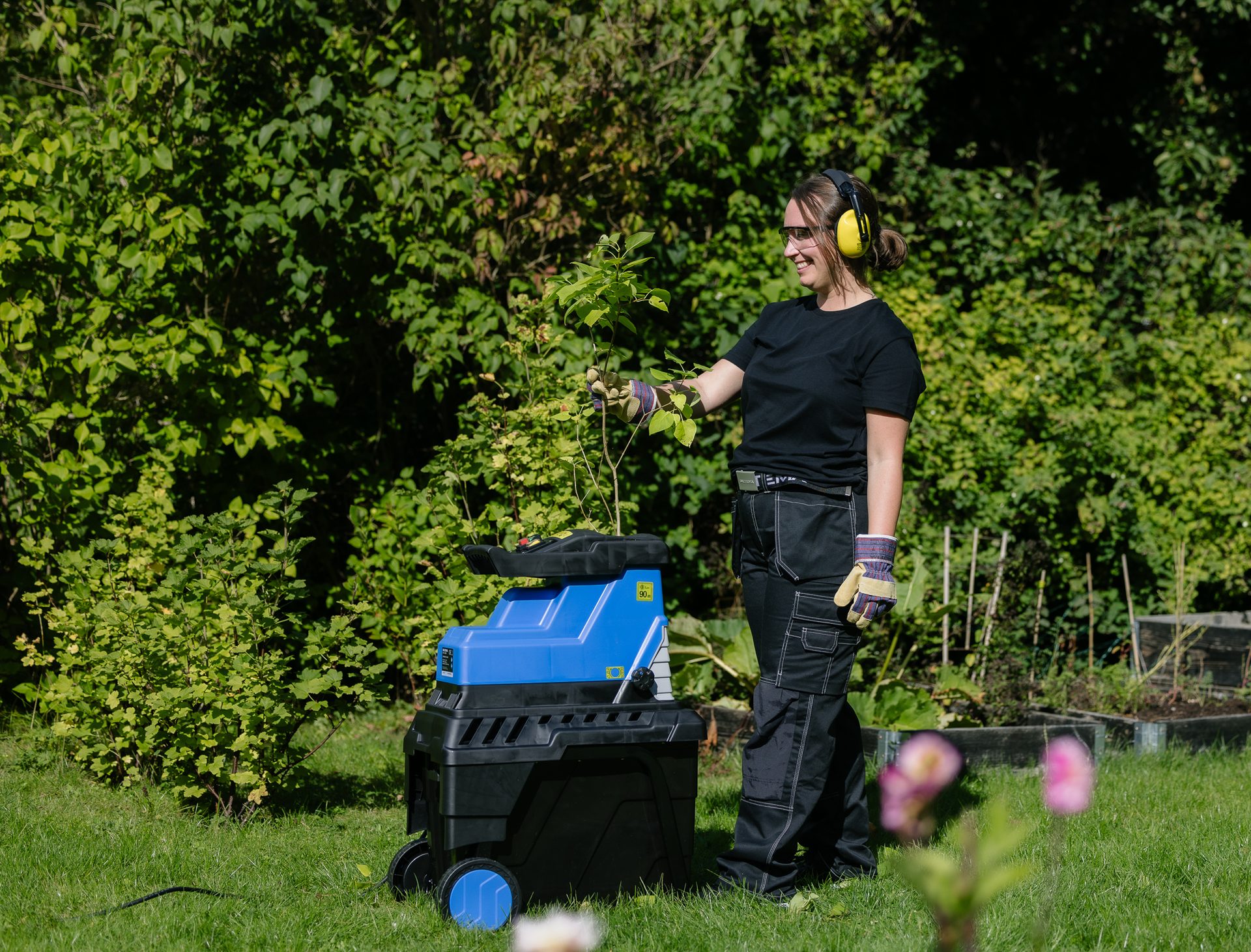 A woman in safety gear feeds branches into a wood chipper in a sunny garden, mulching green waste.