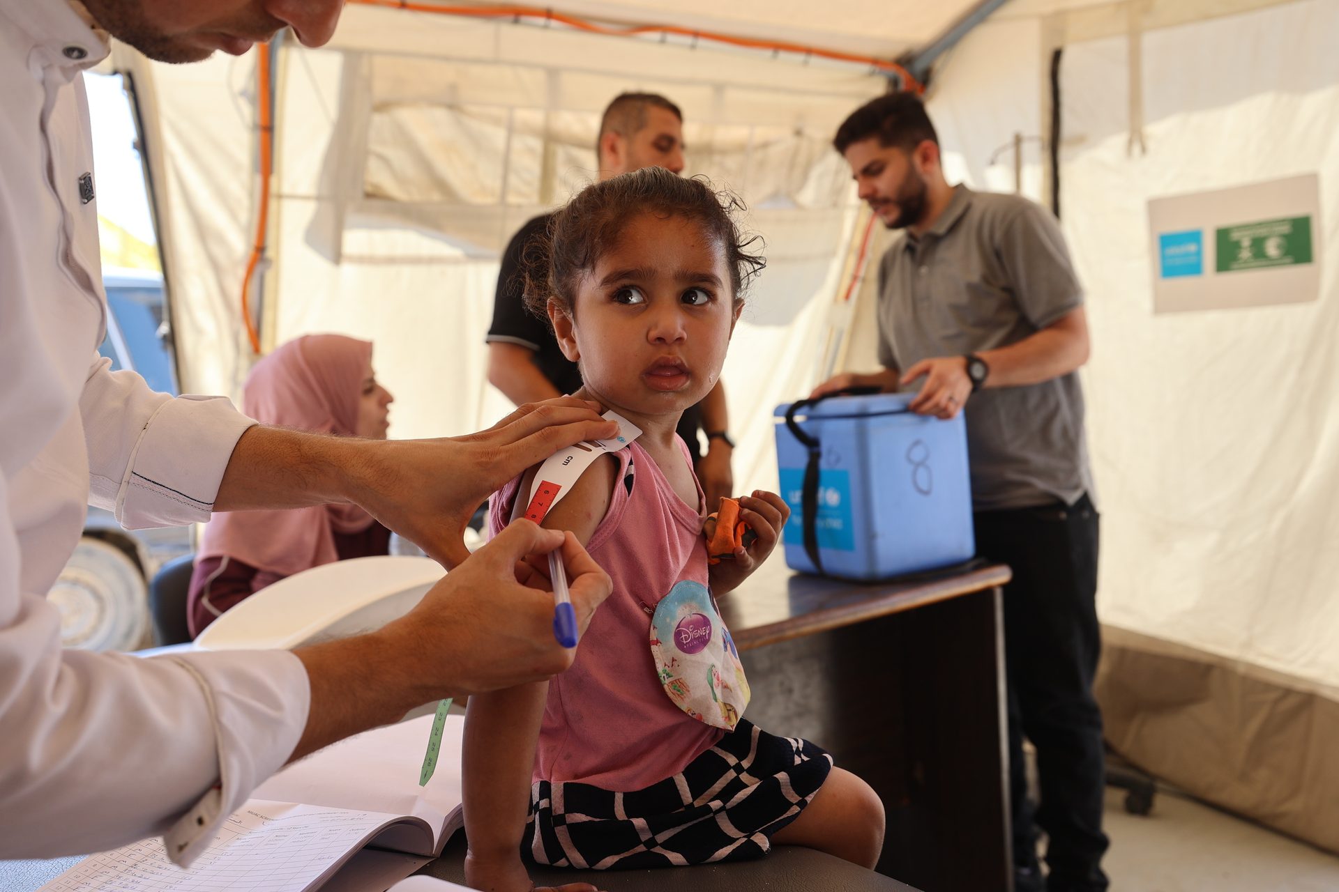A healthcare worker measures a child's arm in a tent, with other people in the background.